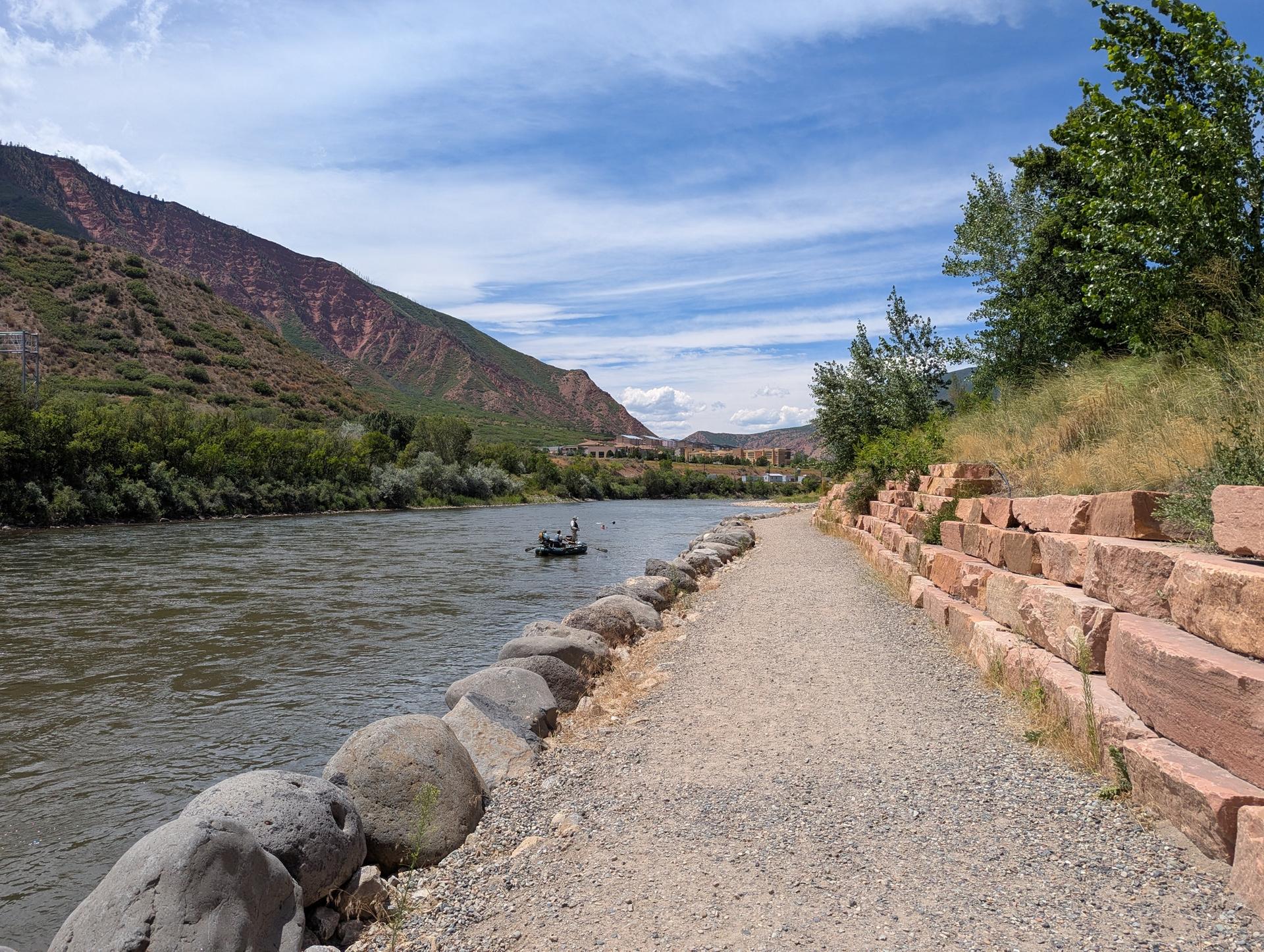 A person in a small boat on a river next to a gravel path and mountains.