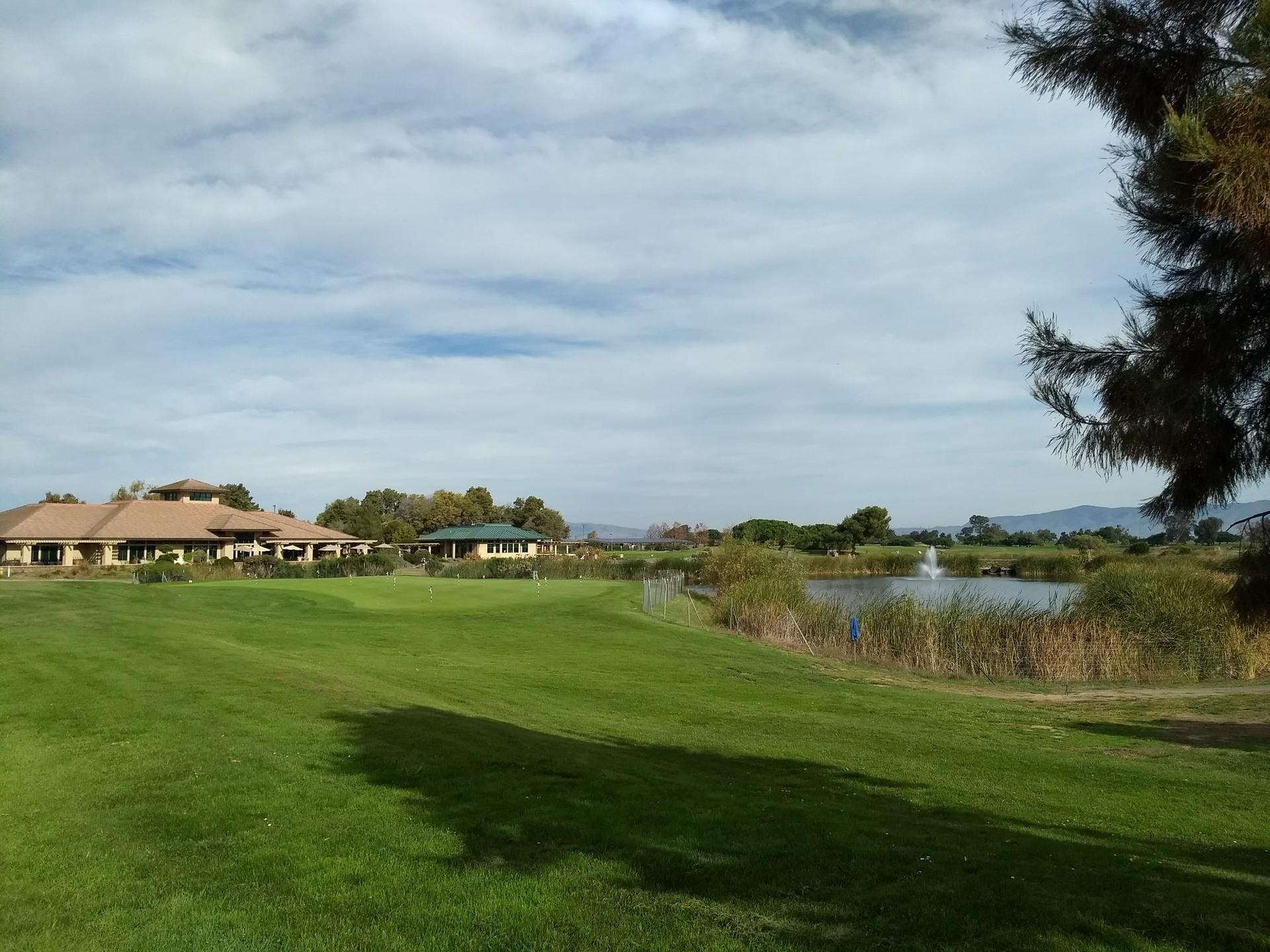 Green golf course with buildings, a pond, and a fountain under a cloudy sky.