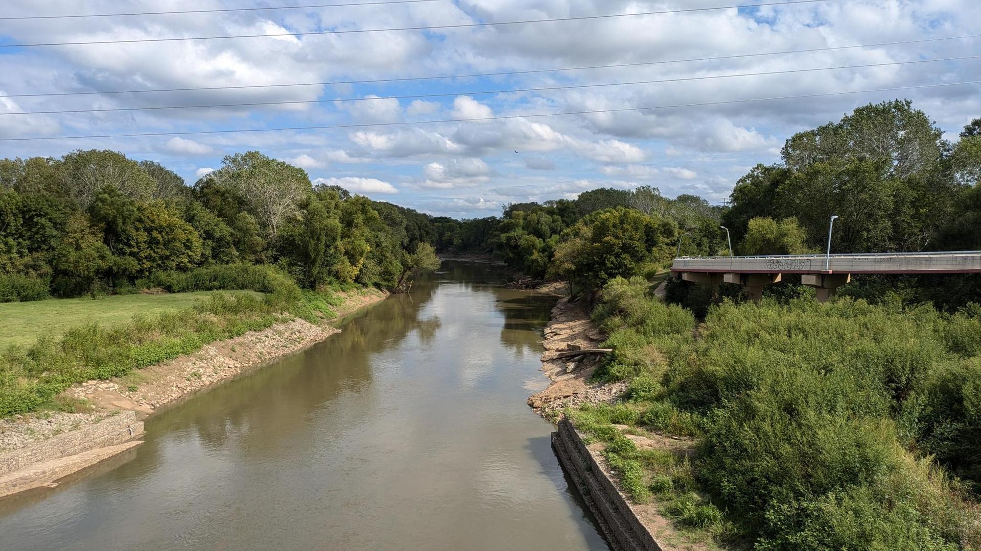 A wide river flows under a bridge, surrounded by lush green trees under a cloudy sky.