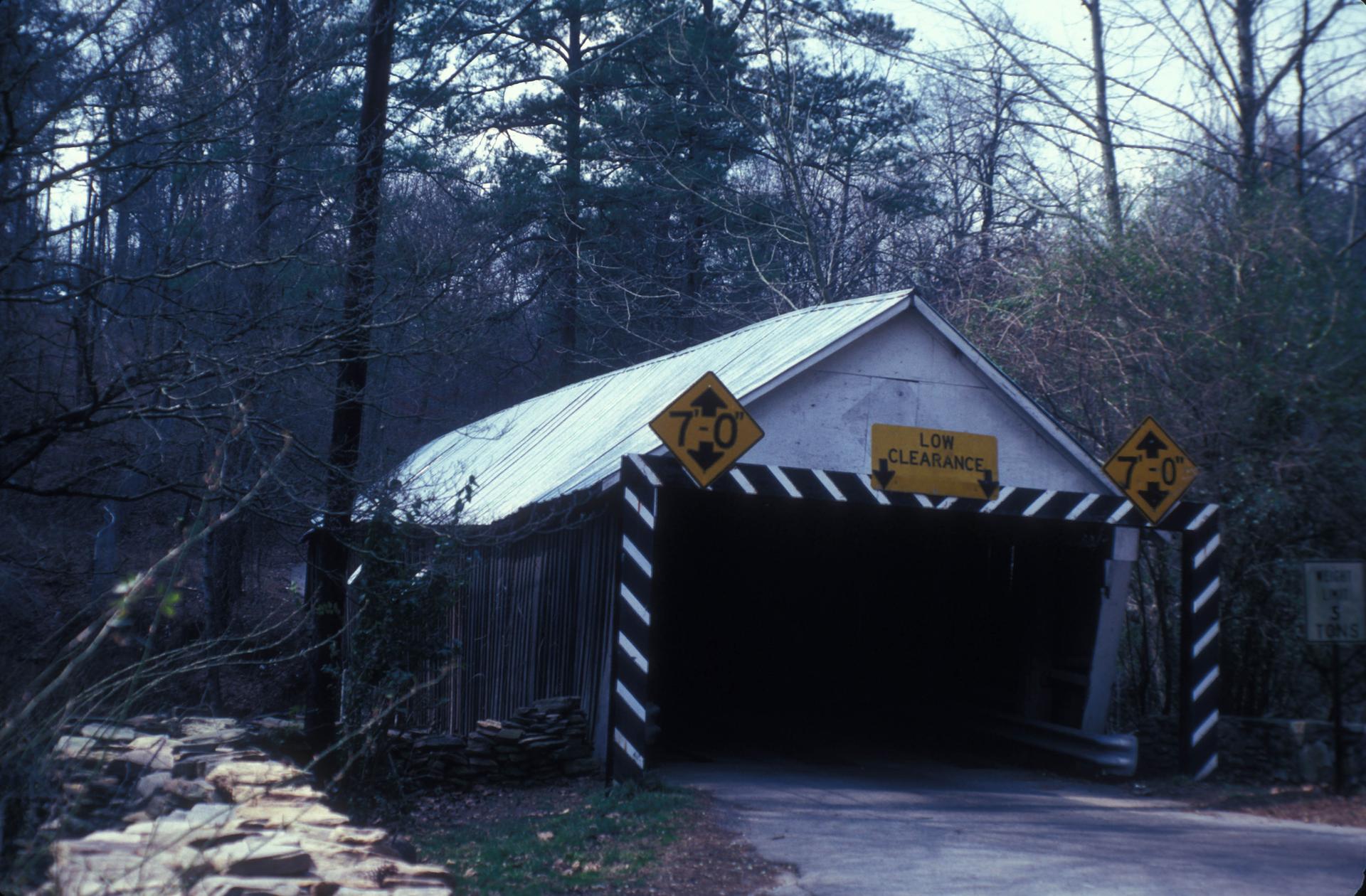 A dark covered bridge with low clearance signs in a dense forest.
