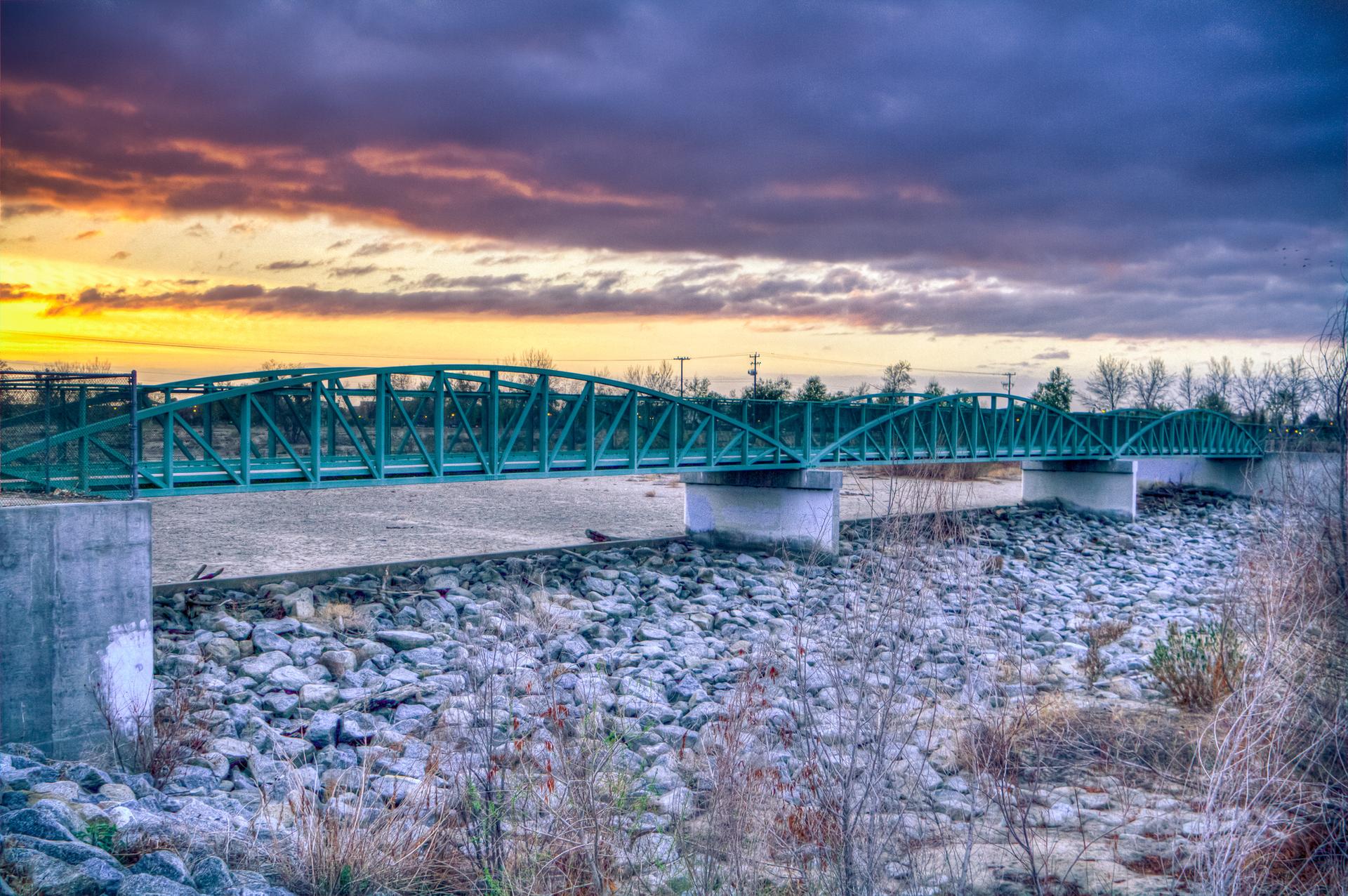 A green pedestrian bridge spans a rocky riverbed under a colorful sunset sky.