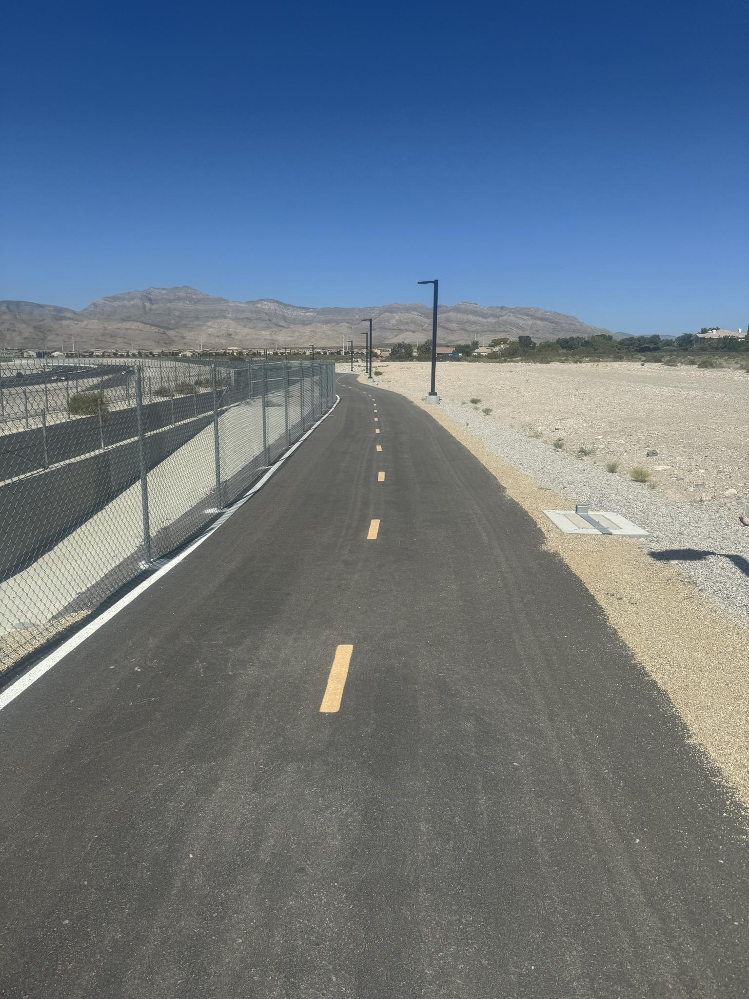Paved desert path with a fence, mountains, and clear blue sky.