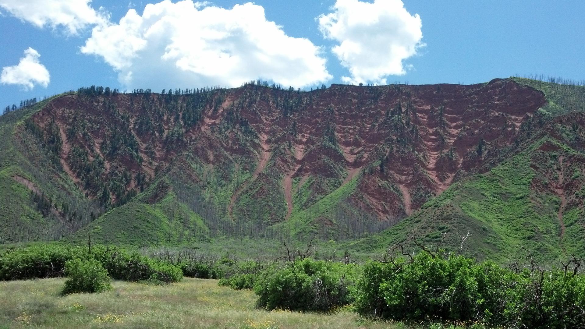 A large reddish-brown mountain with green slopes under a bright, cloudy sky.