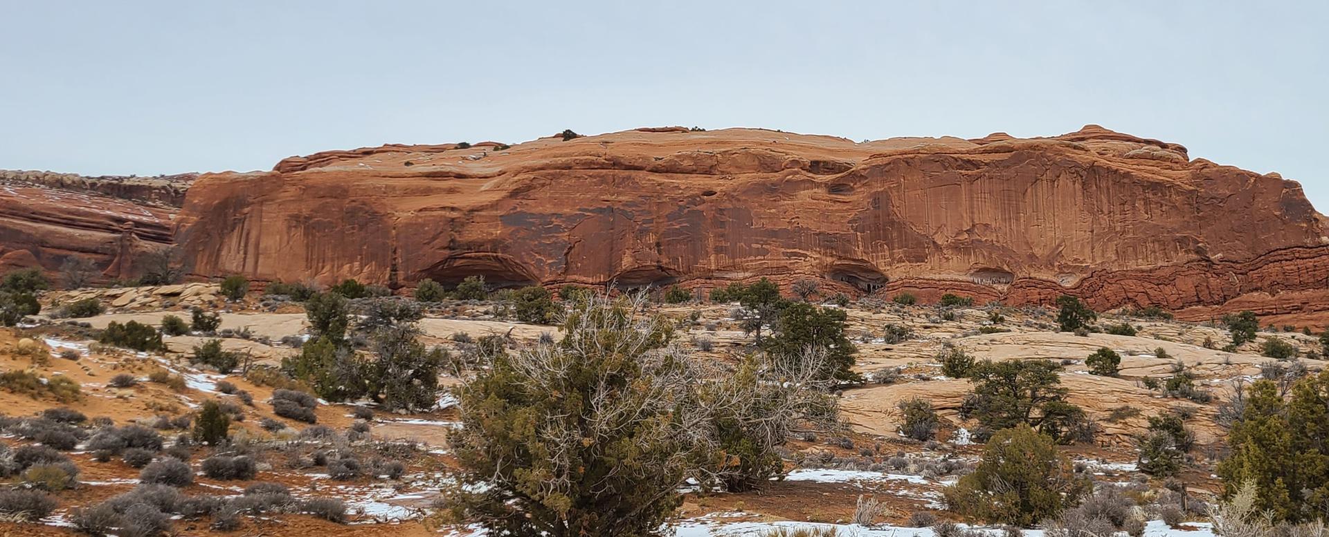Large red rock formation dominates a desert landscape under an overcast sky.