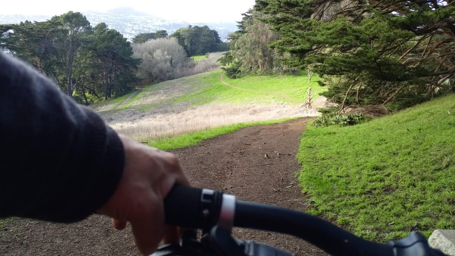 Cyclist's hand on handlebars, riding a dirt path through green hills. - Philosopher's Way