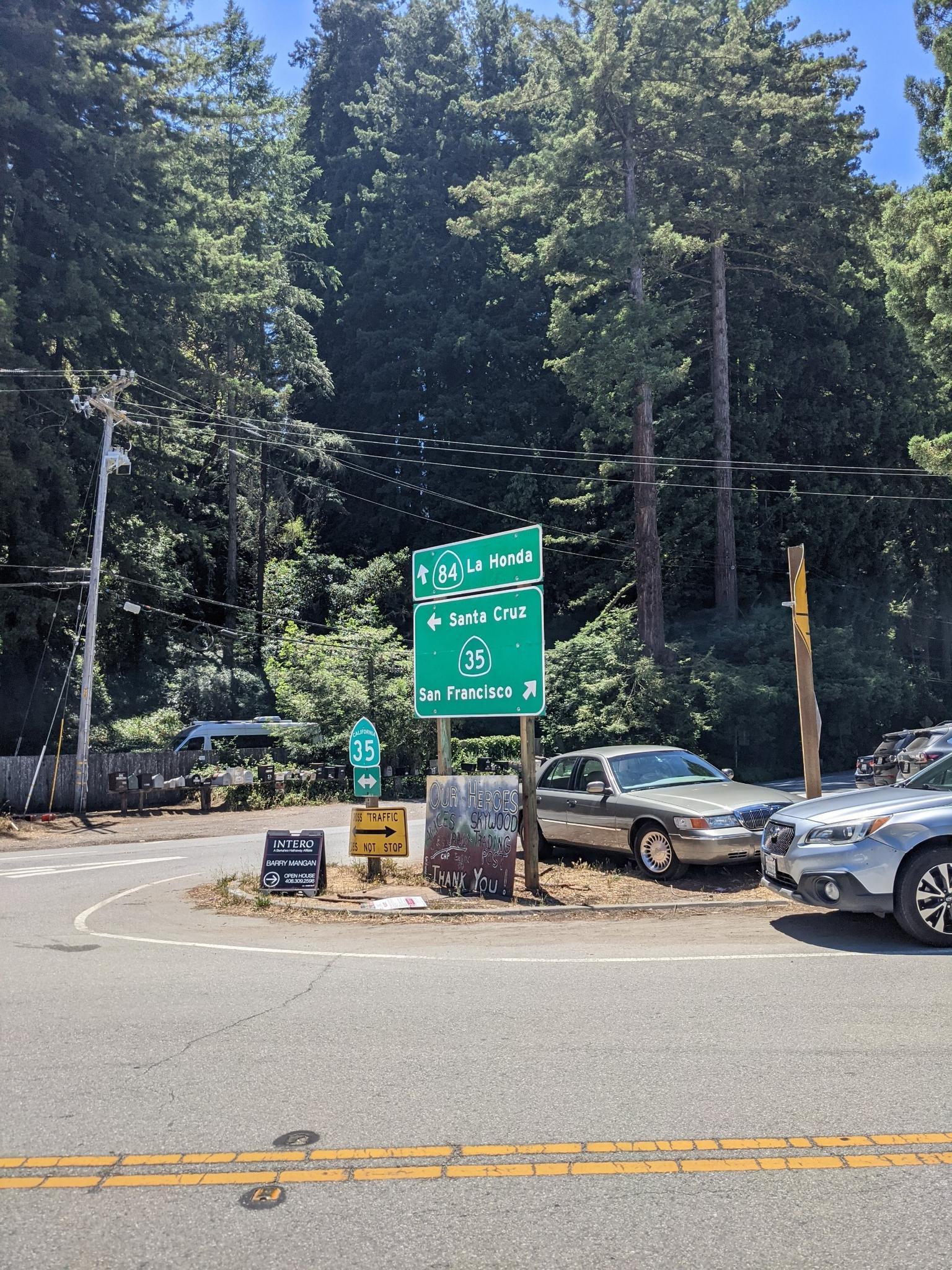 Road signs at an intersection with cars and tall evergreen trees.