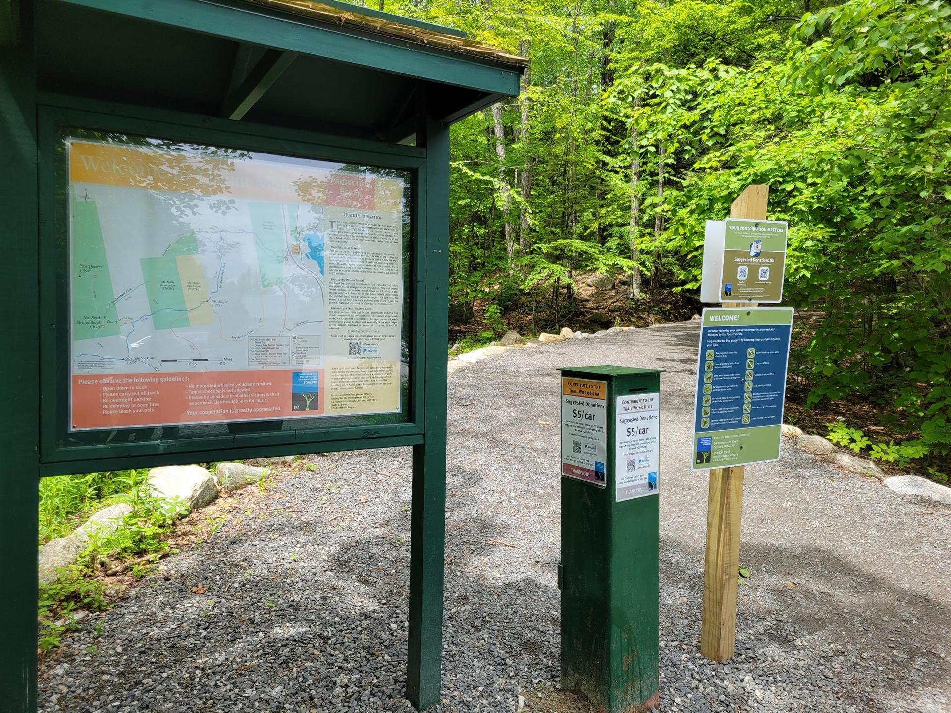 Trailhead sign and information kiosk on a gravel path in a green forest.