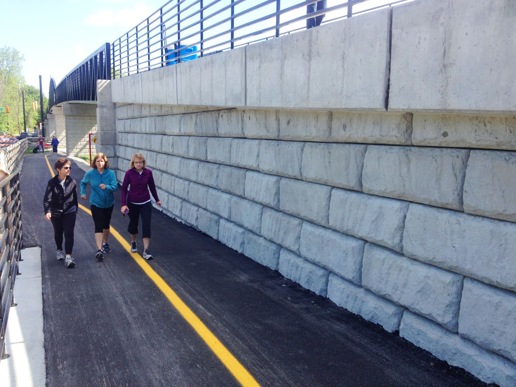 Three women walk on a paved path next to a large concrete retaining wall. - Monon Trail North Trailhead – Monon Trail loop from Sheridan