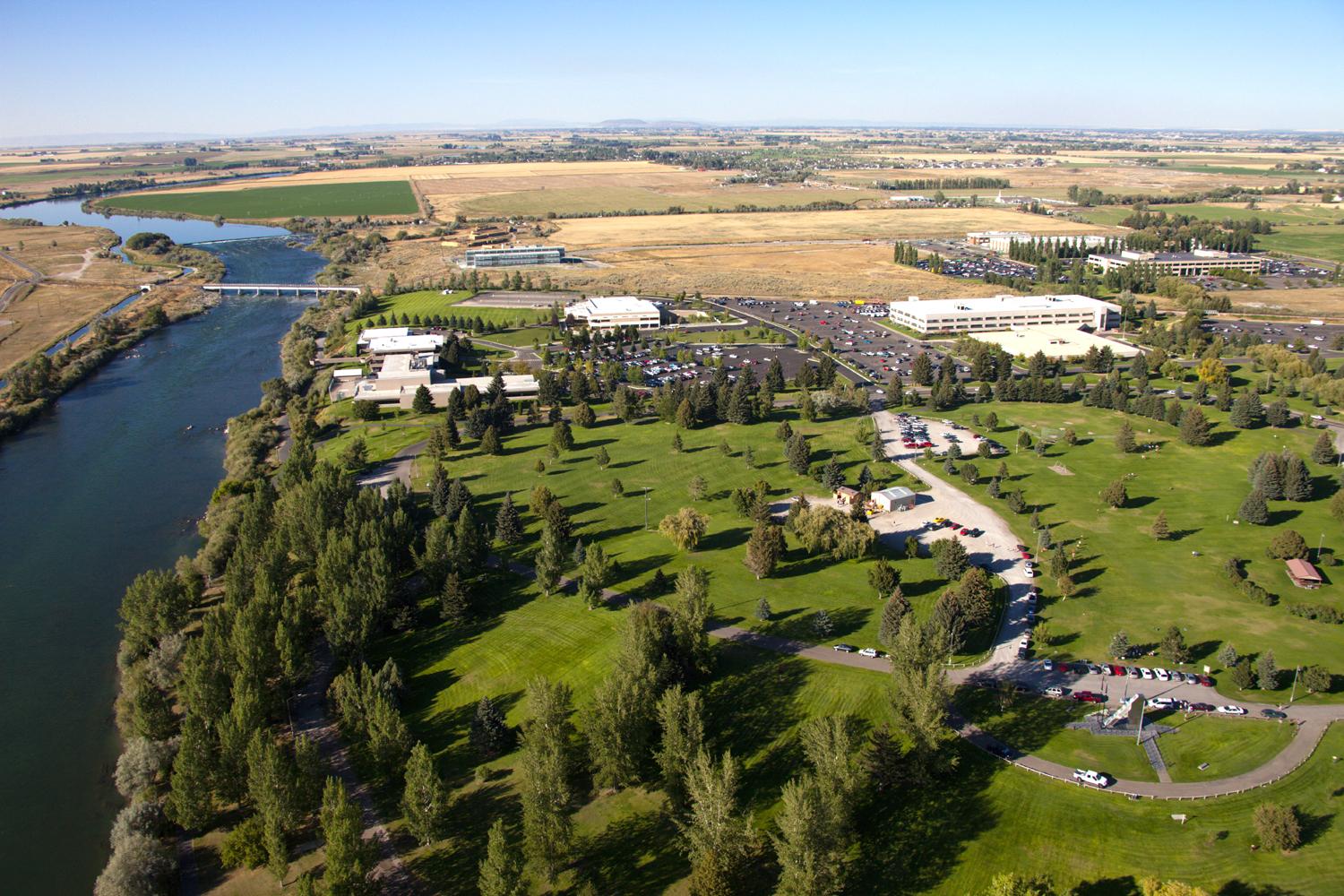 Aerial view of a river flowing past green fields, buildings, and parking lots.