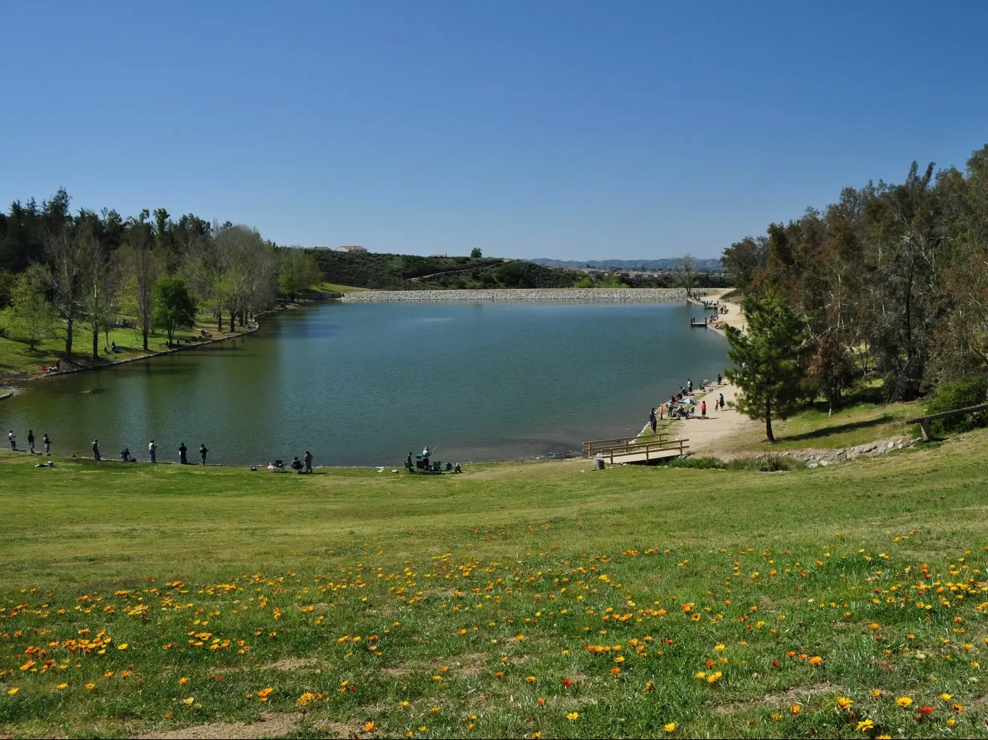 A wide lake with many people fishing along its grassy shores on a sunny day.