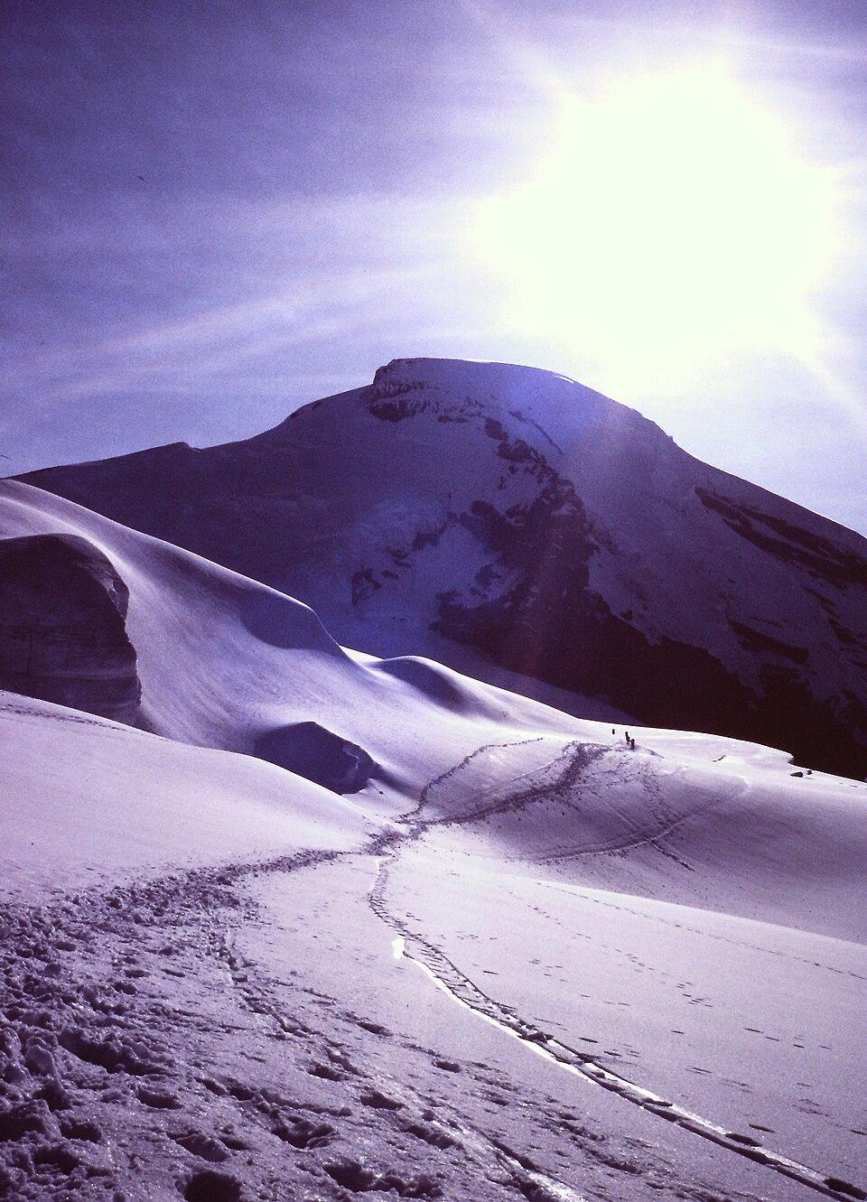 Bright sun over a vast, snow-covered mountain with a winding trail.