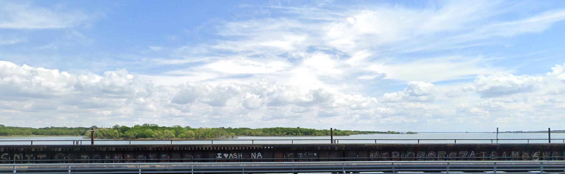 Wide water view over a bridge, with distant trees under a cloudy sky.