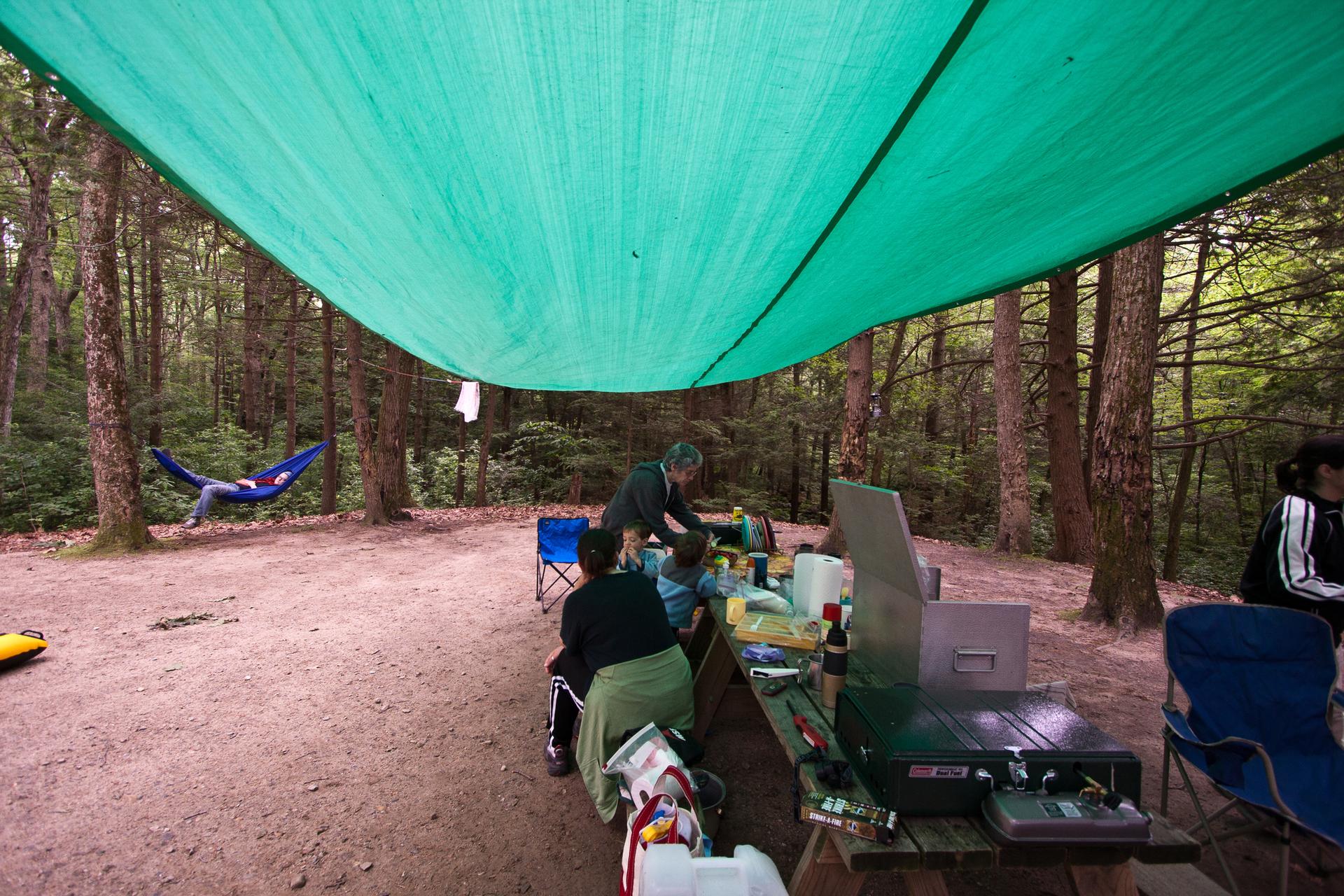 People cooking and relaxing at a shaded forest campsite under a green tarp.