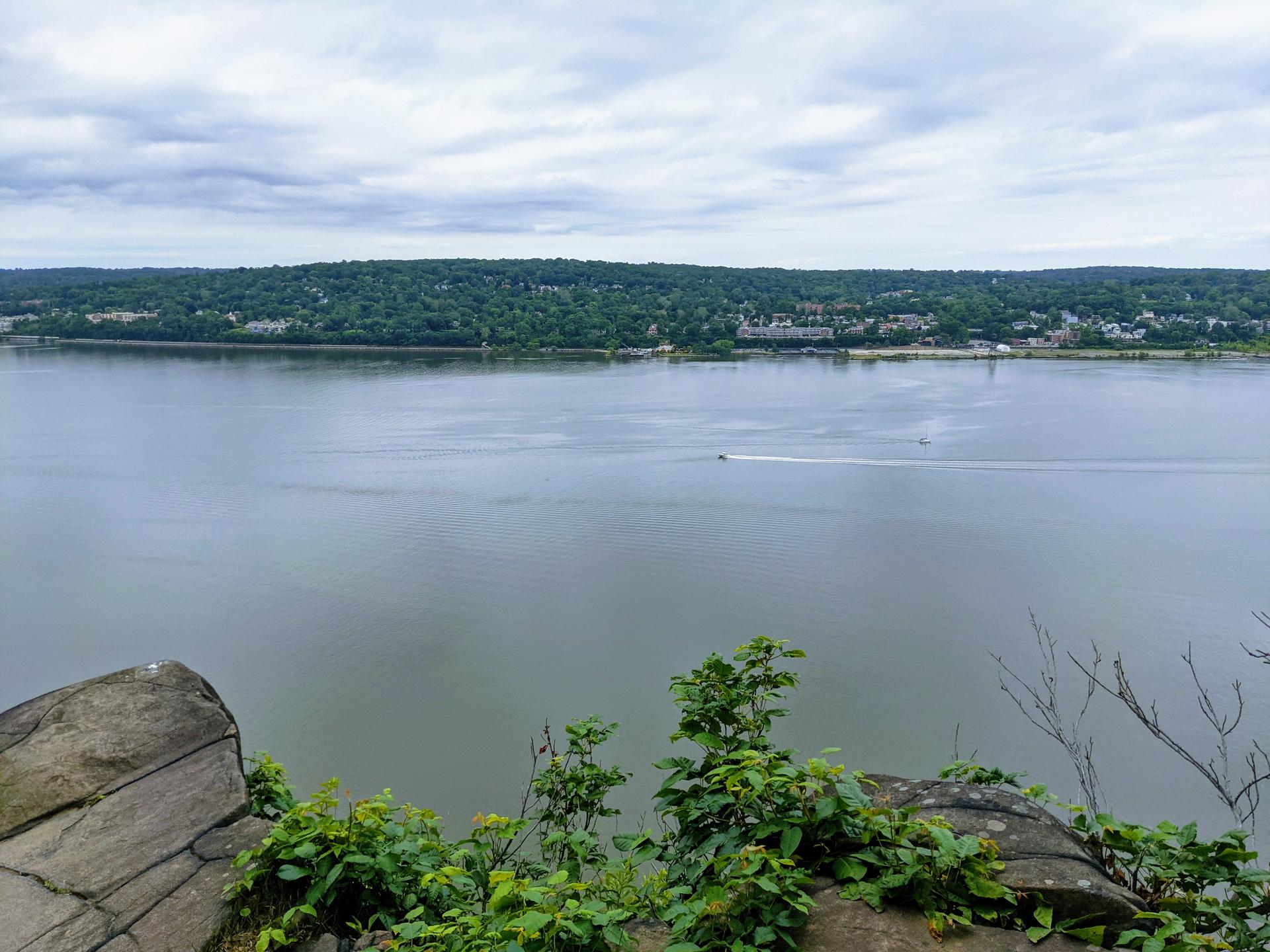 Wide river with a small boat, distant forested shore, under cloudy skies.