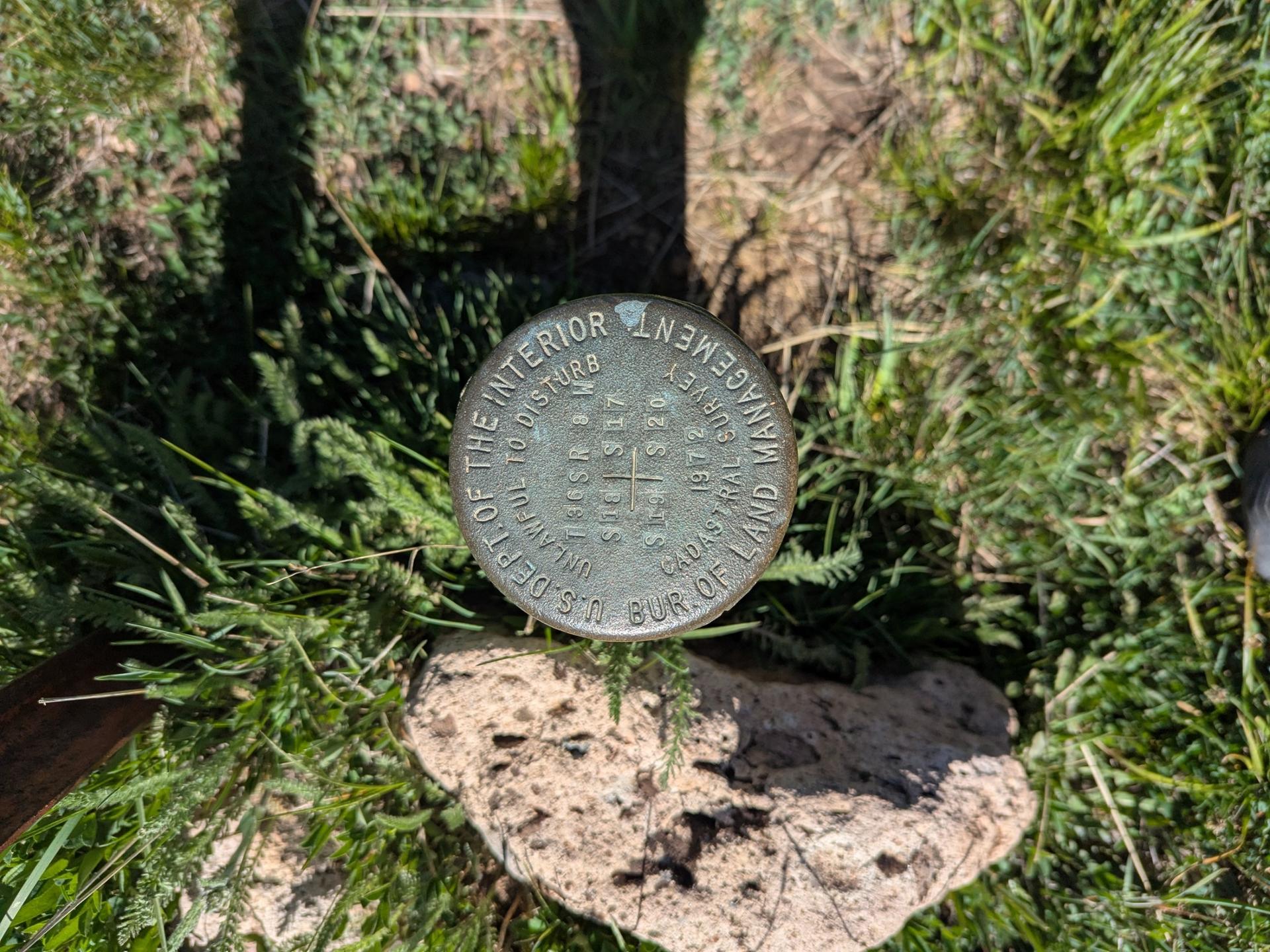 A metal survey marker embedded in grassy ground with a rock, under sunlight.