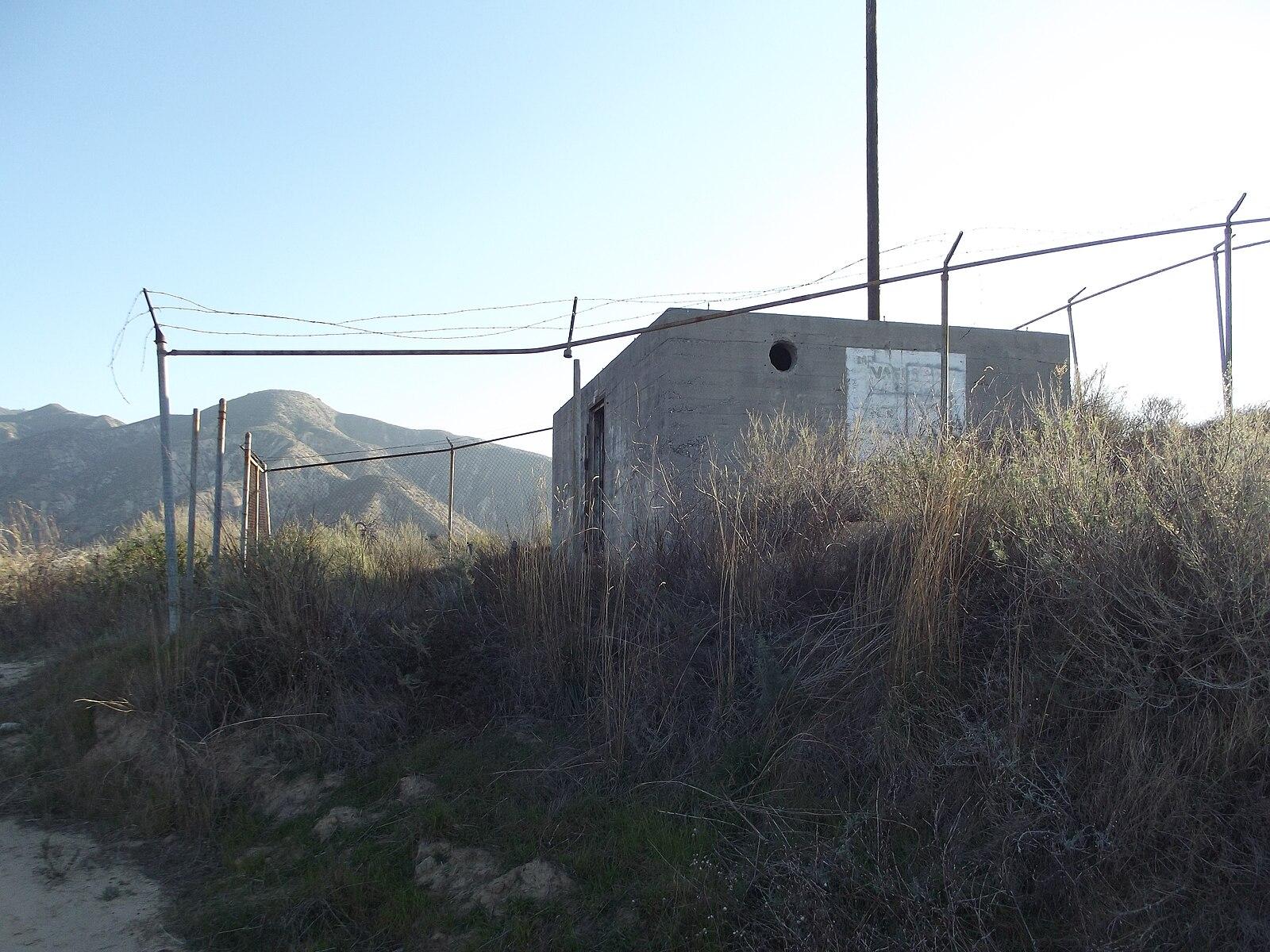 Concrete building behind a fence on a dry hill with mountains in background.