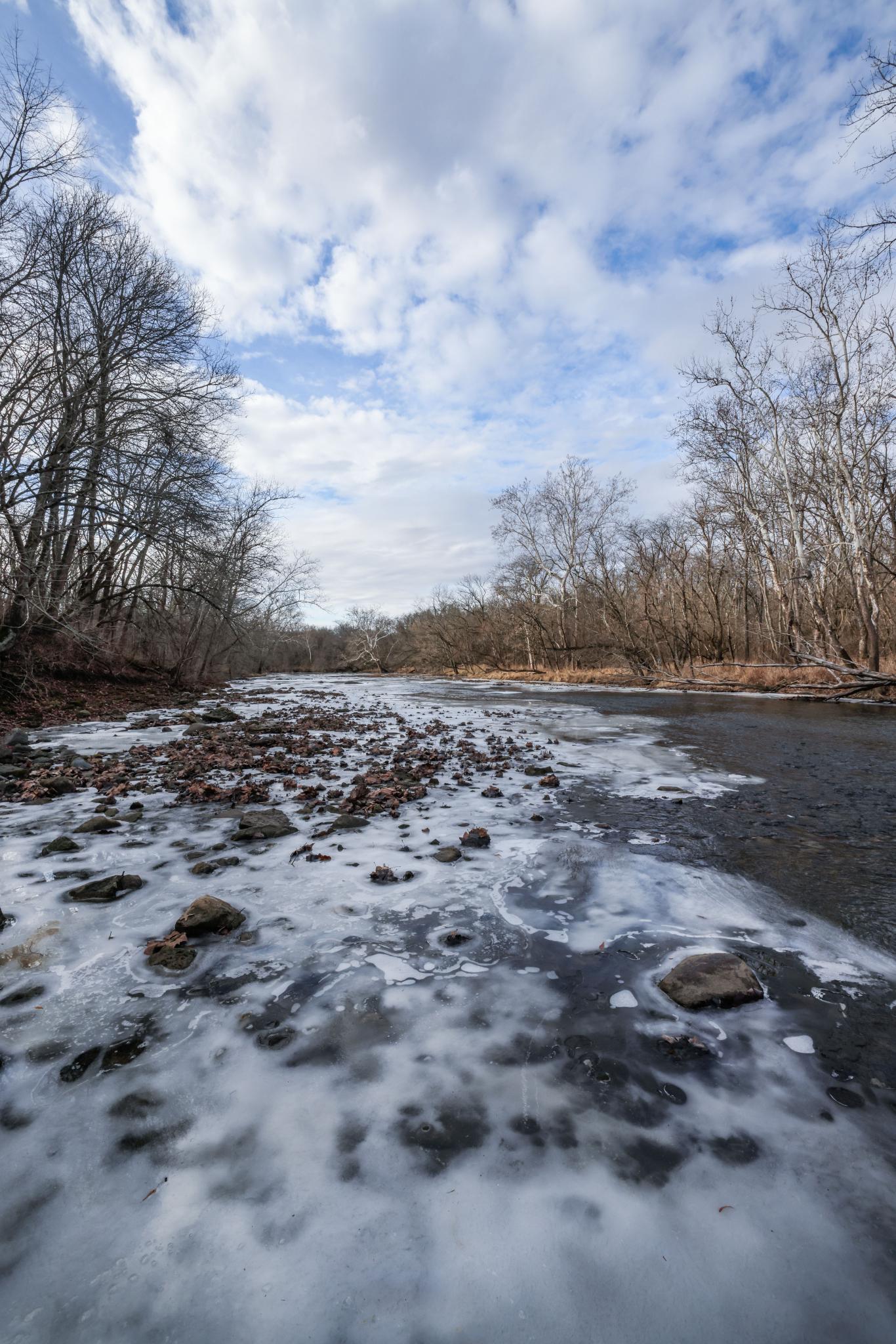 Icy riverbed with bare trees under a bright, cloudy winter sky.