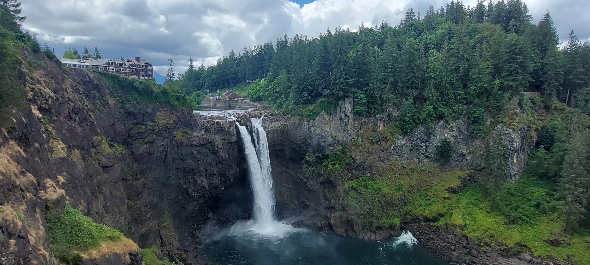A powerful waterfall cascades into a pool, with a building and forest above.