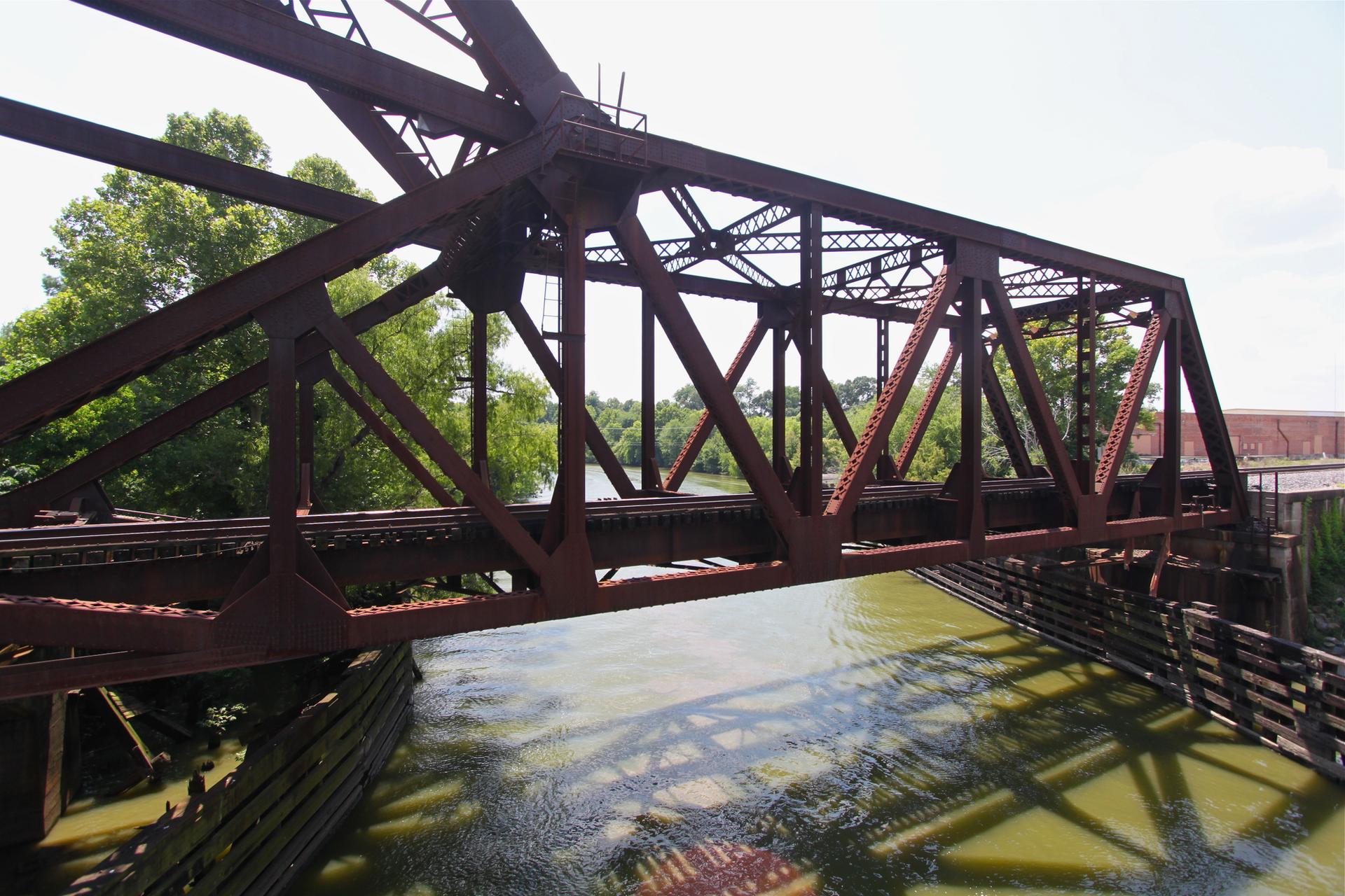 Rusty red truss bridge spans a river with green trees under bright sun.