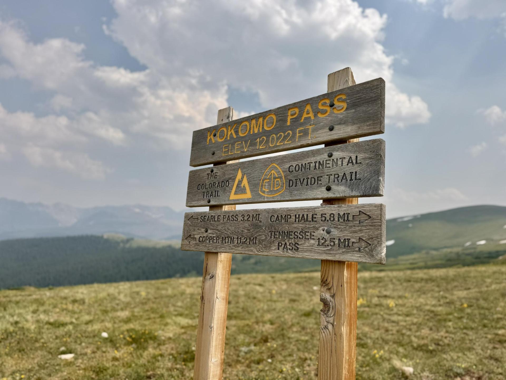 A wooden trail sign stands on a grassy mountain with distant peaks.