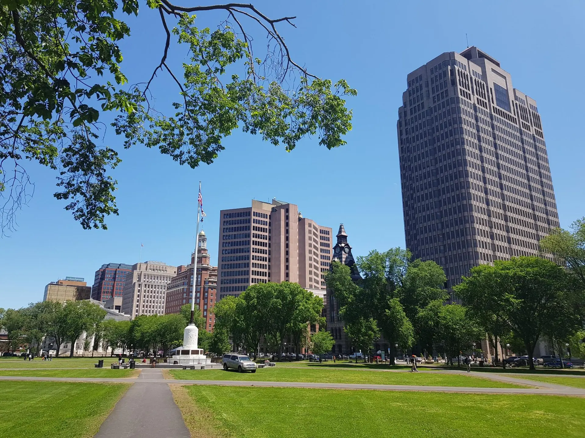 City park with tall buildings, green grass, and people on a sunny day.