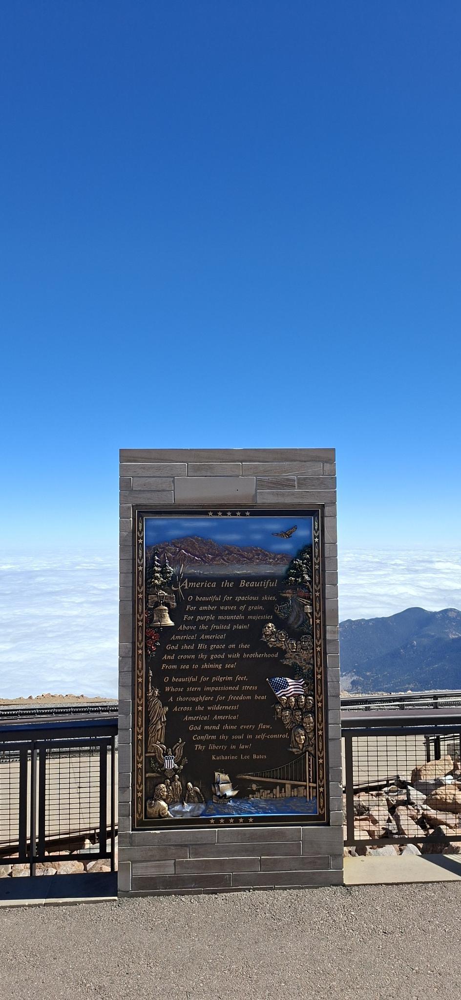 Informational sign on a mountain overlook, with clouds below and blue sky.