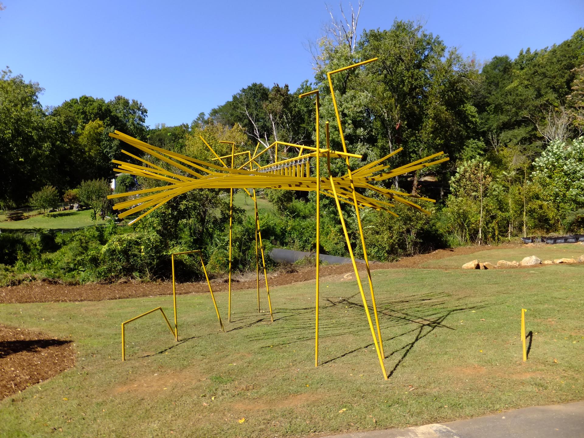 Large yellow abstract sculpture on a sunny grassy field with green trees.