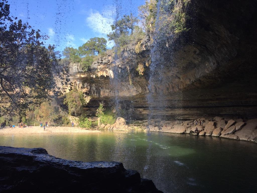 View from behind a waterfall into a green pool with people on a sunny bank.