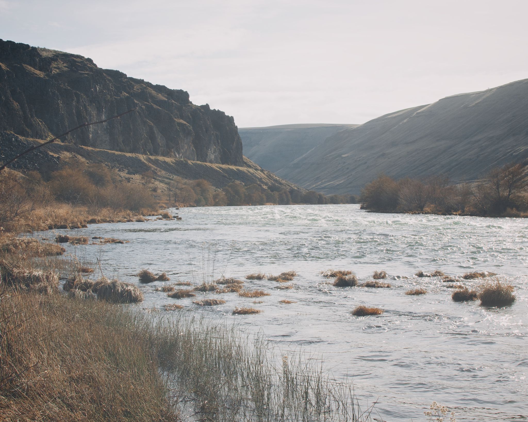 A wide river with rocky banks flows through a sunny, hazy canyon. - Deschutes River Trail