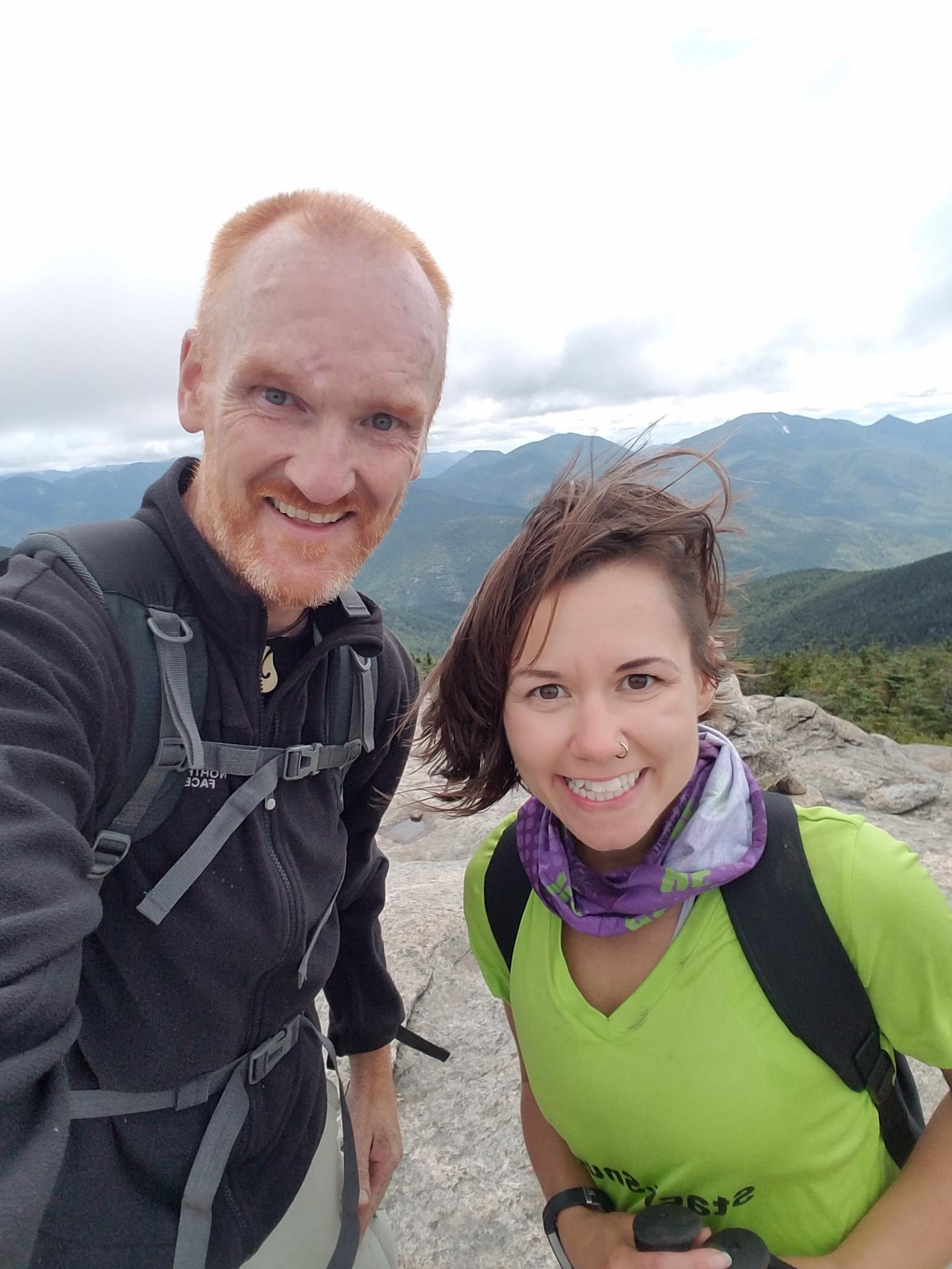 Two smiling hikers pose for a selfie on a windy mountain peak.