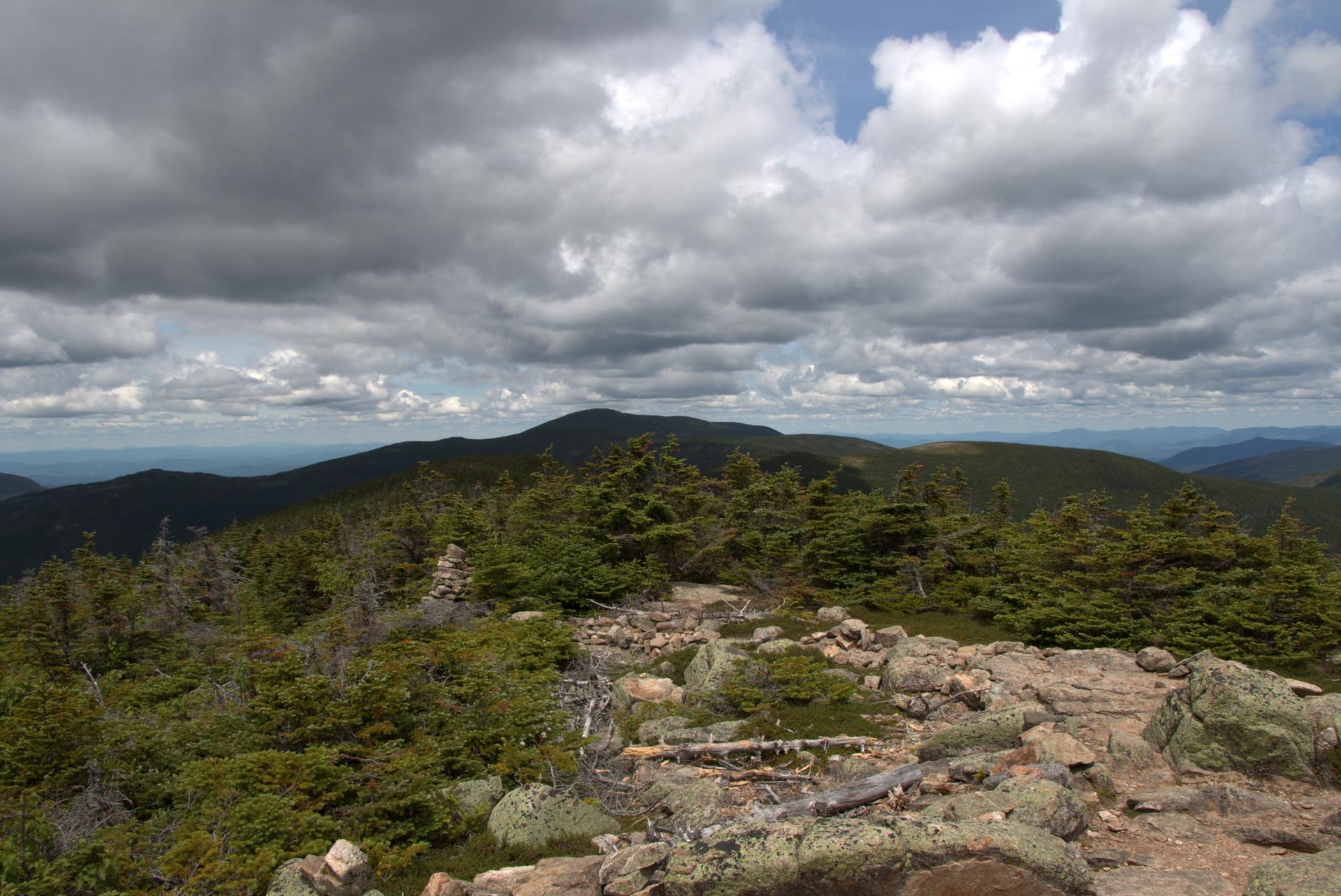 Rocky mountain terrain with evergreen trees under a dramatic cloudy sky.