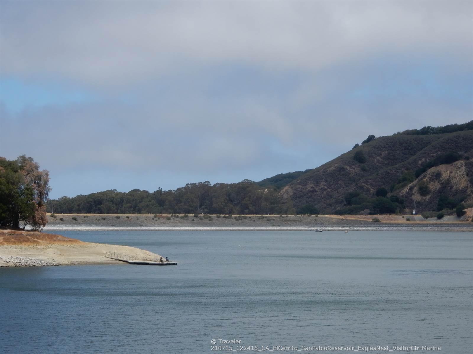 Wide lake with a rocky shore and distant hills under a cloudy sky.