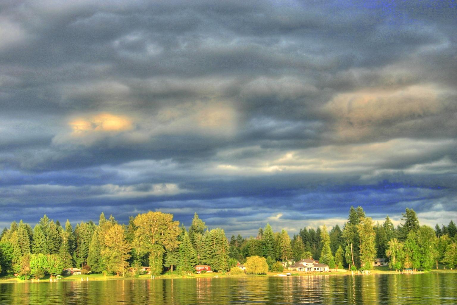 Lake shore with houses and trees under a dramatic, cloudy sky.