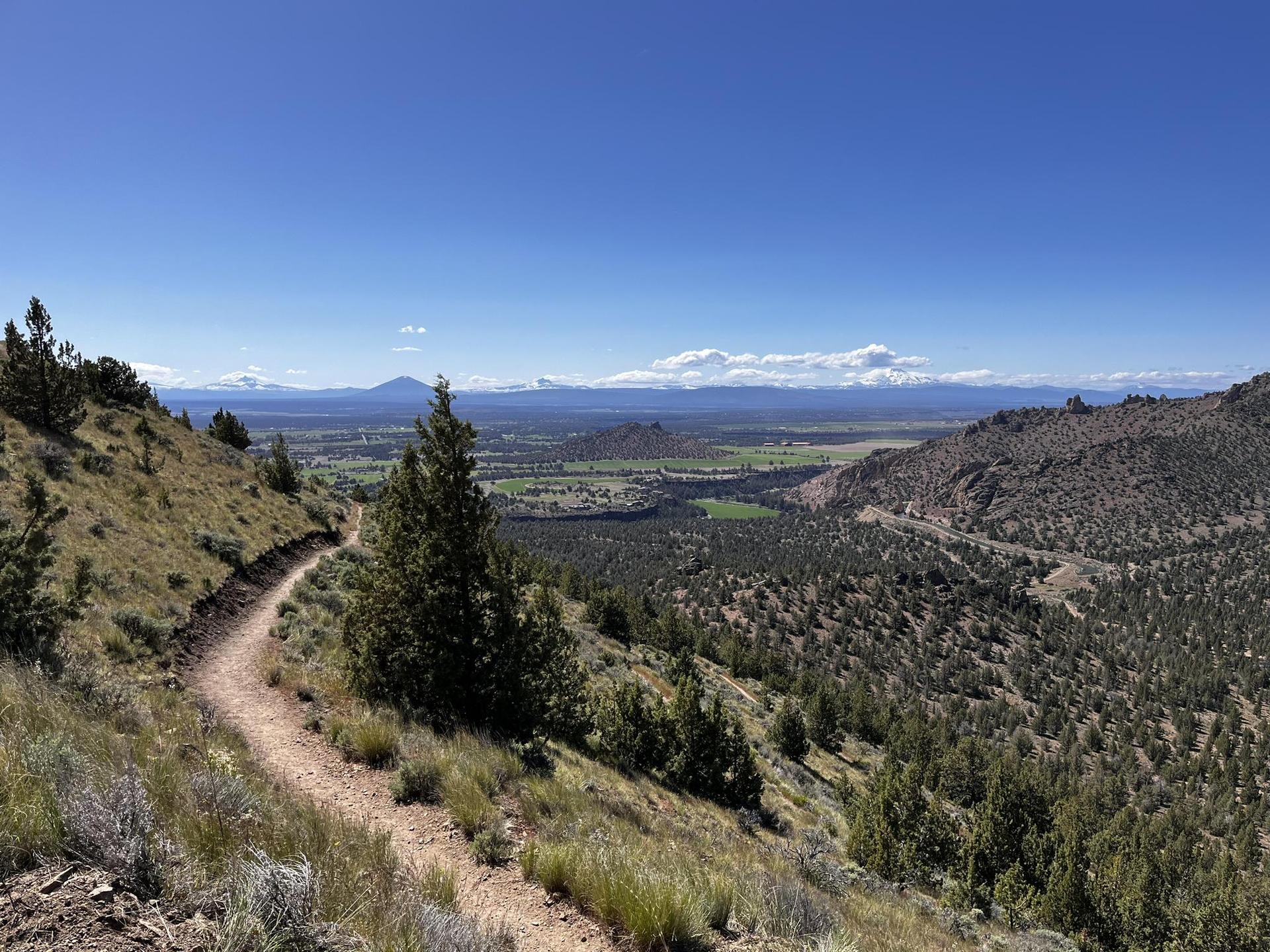 Winding dirt trail on a sunny hillside overlooking a wide valley and mountains.