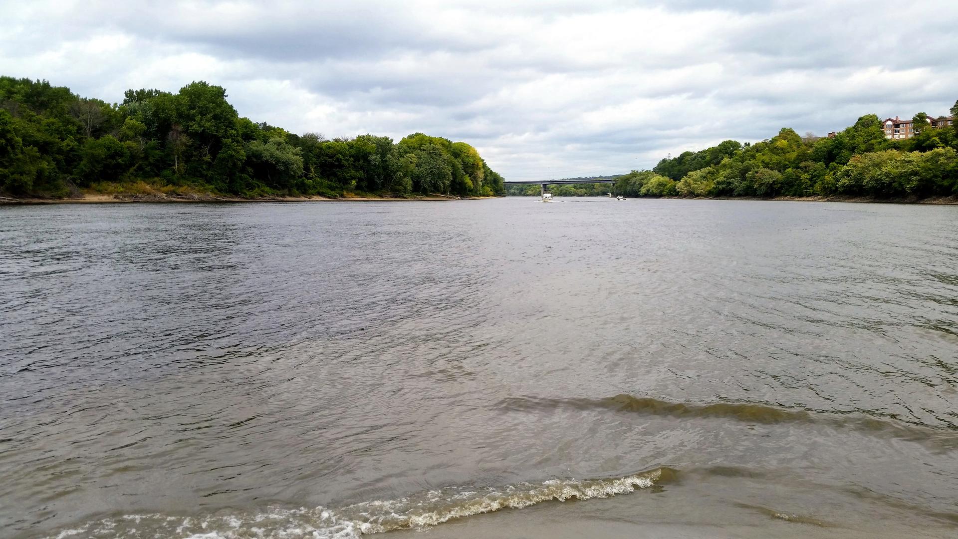 Wide river with small waves, surrounded by green trees under a cloudy sky.