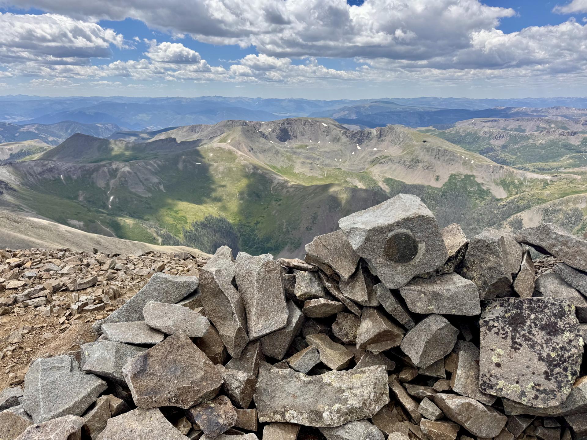 A rock cairn on a mountain peak overlooking a vast, cloudy landscape.
