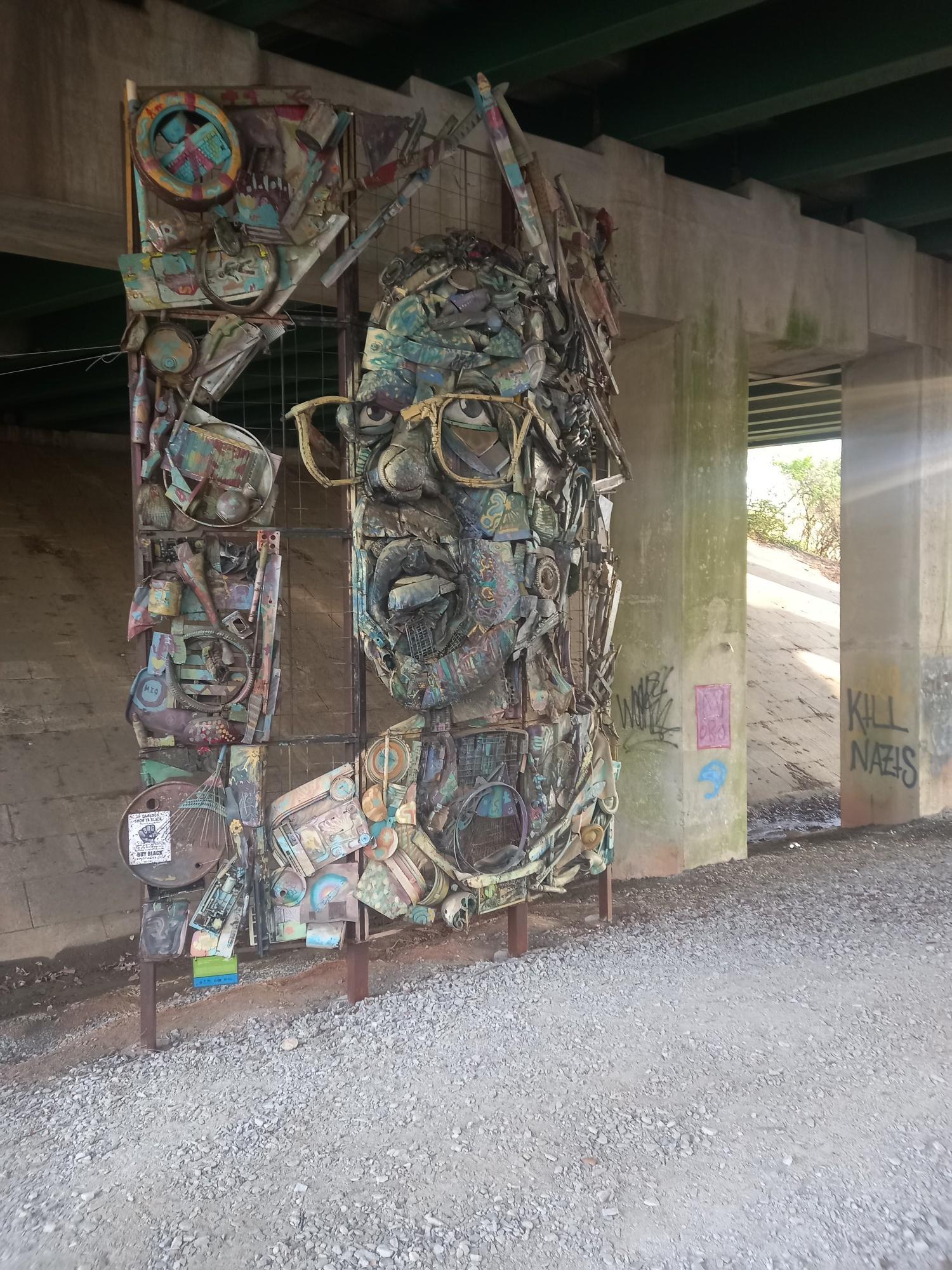 A large, colorful, abstract sculpture of a face with glasses under a bridge.