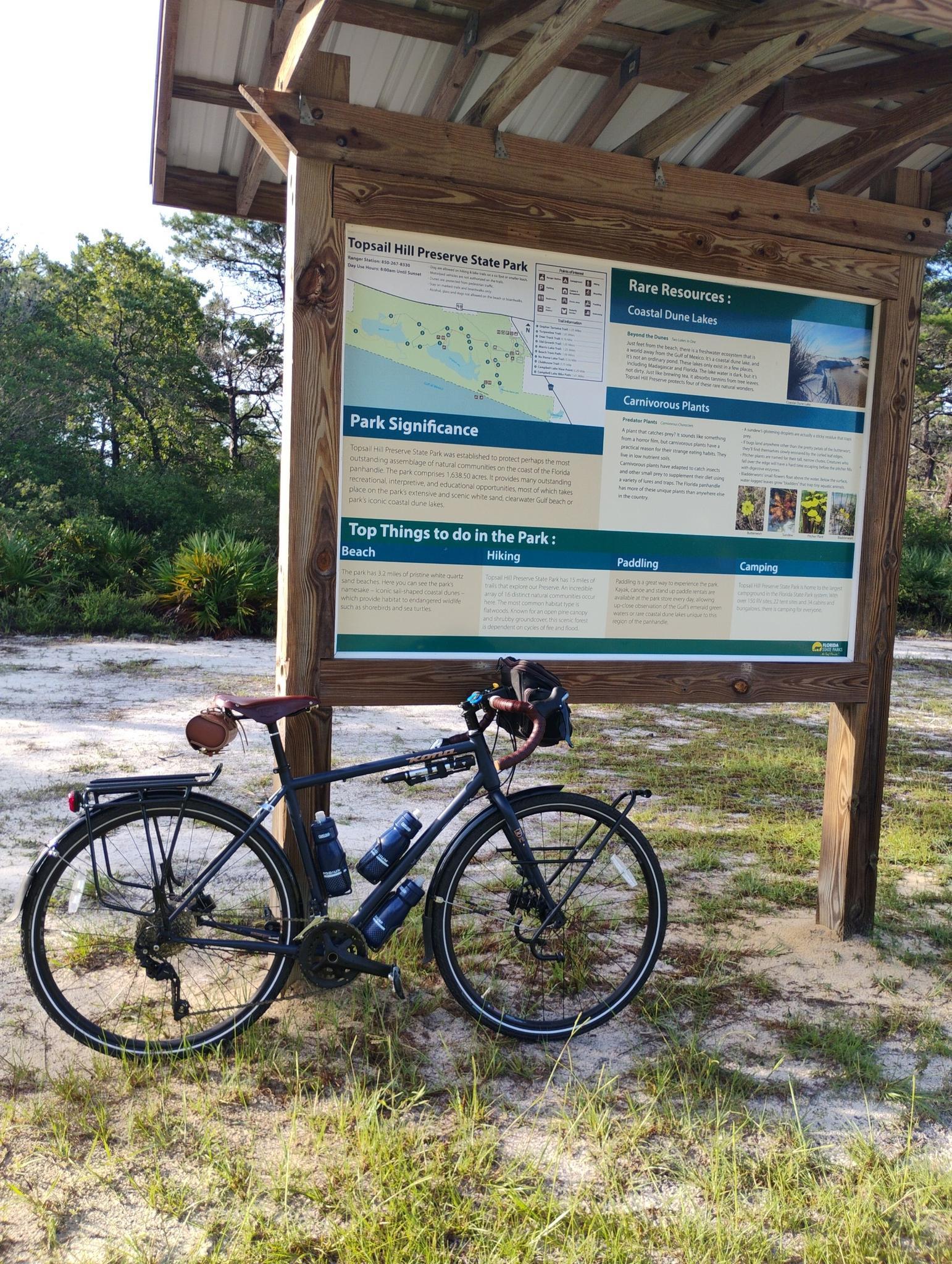 A bicycle is parked on sandy ground next to a large outdoor information sign.