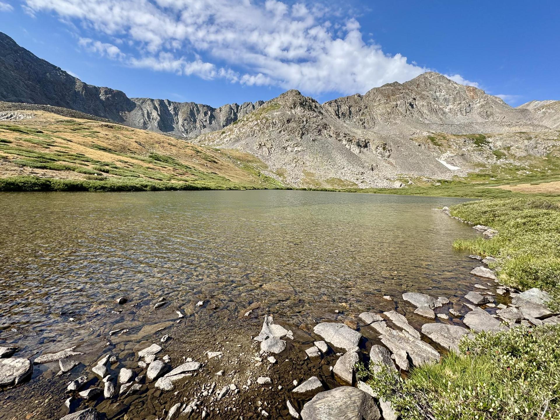 Clear mountain lake with rocky shore, rugged mountains under a blue sky.