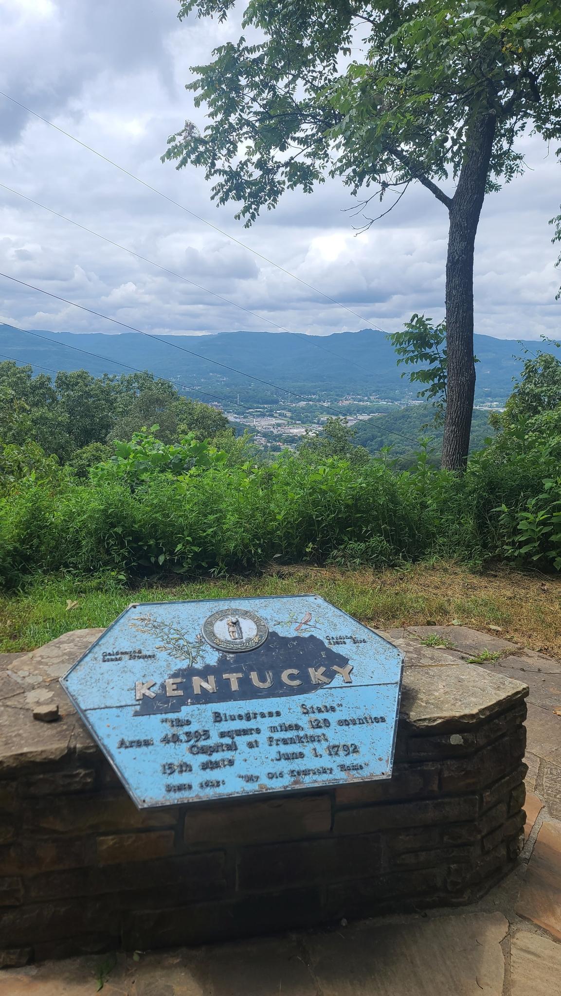 Blue hexagonal sign on stone base, overlooking a town and mountains under cloudy sky.
