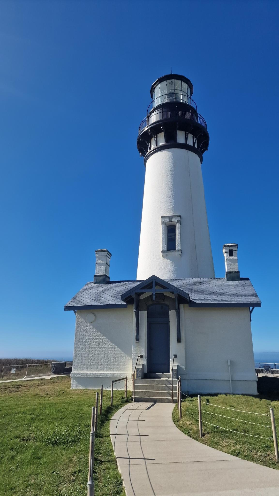 A tall white lighthouse stands against a clear blue sky with a paved path leading to it.