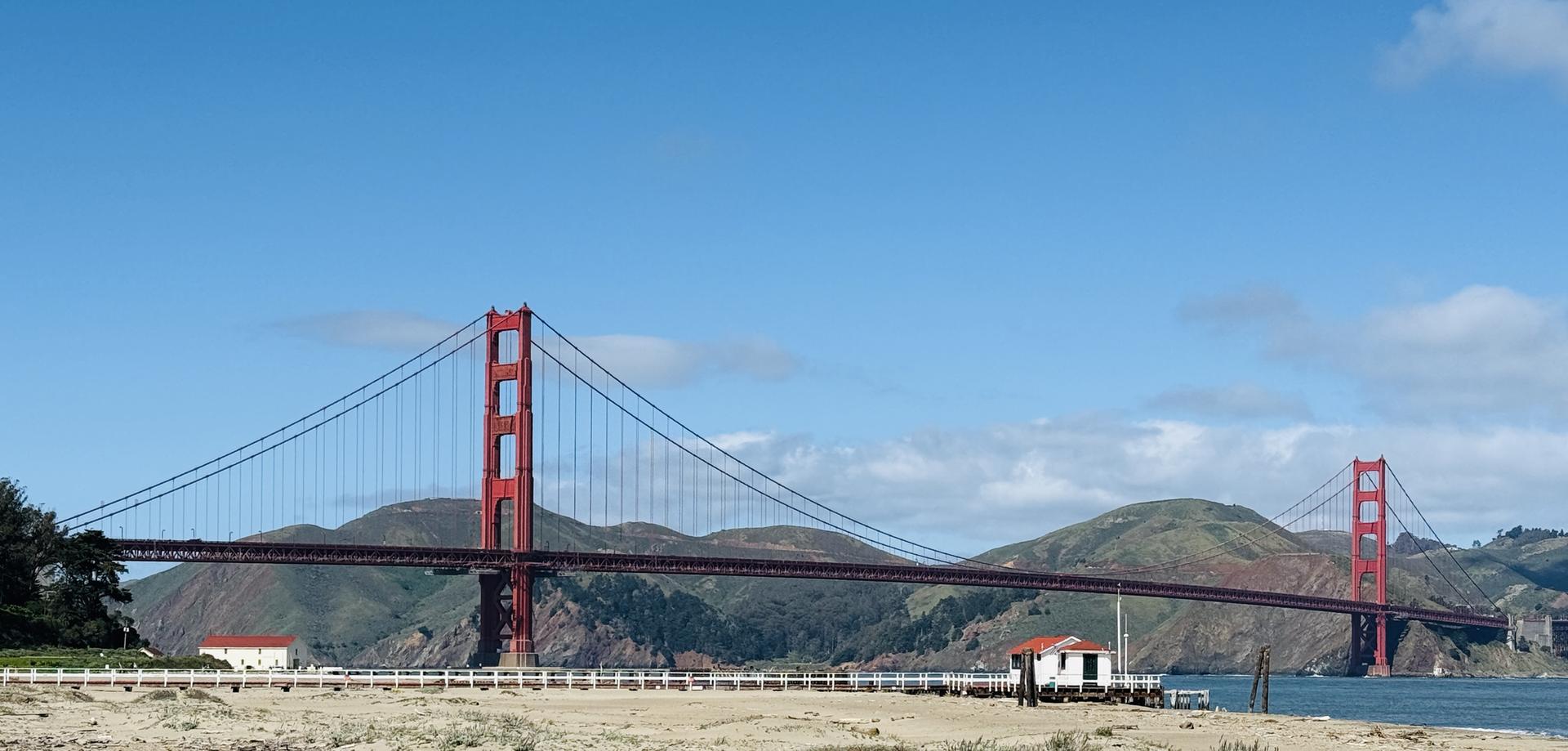 A large red suspension bridge spans water, with hills under a clear sky.