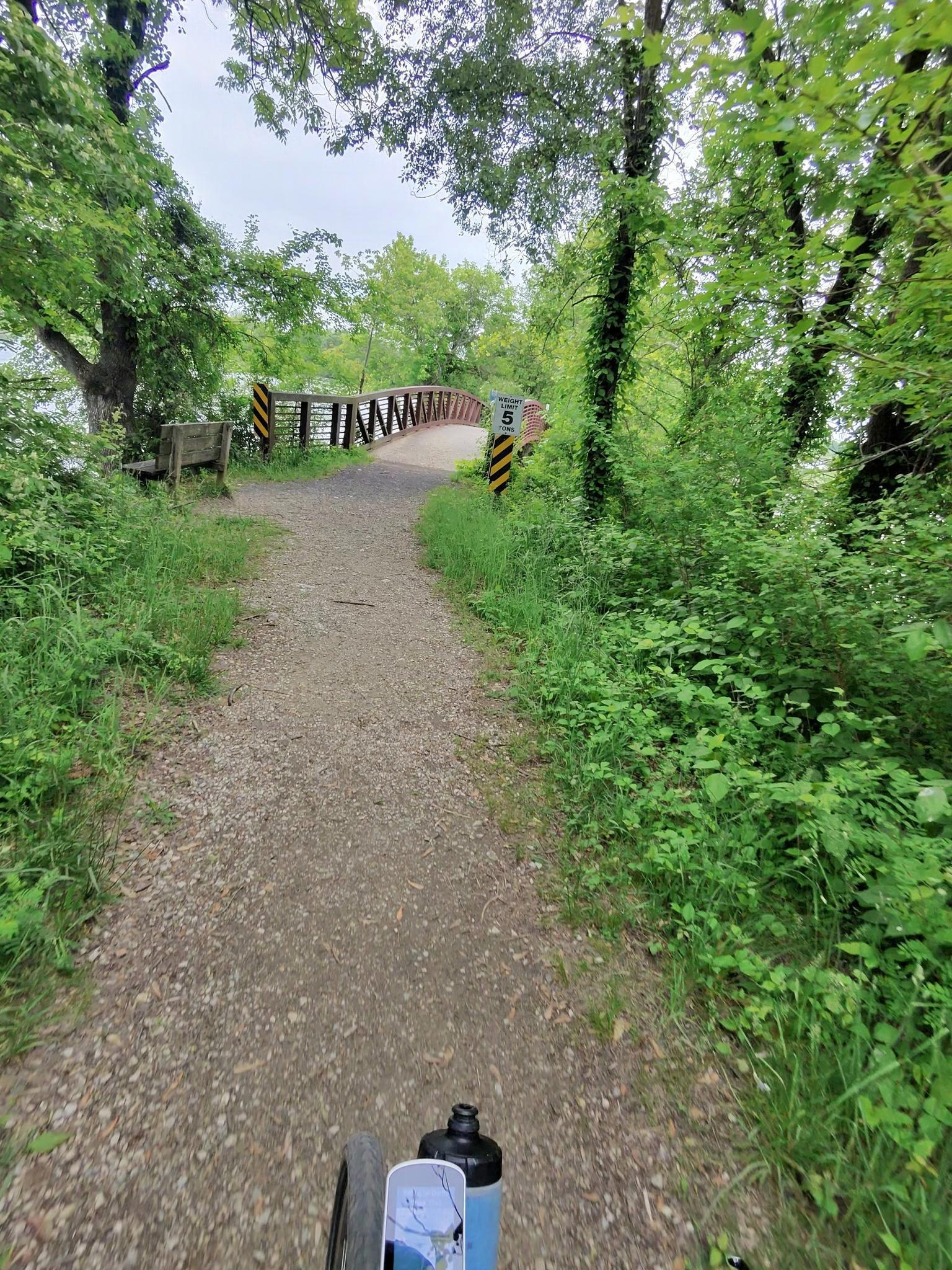 A gravel path leads to a bridge surrounded by dense green trees.