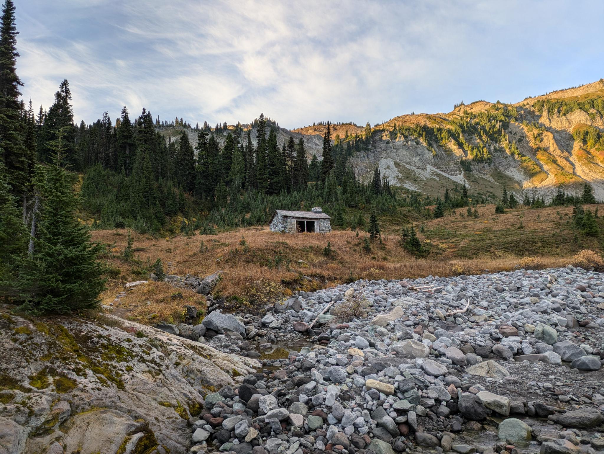 A small rustic cabin in a grassy field with a rocky riverbed and mountains.
