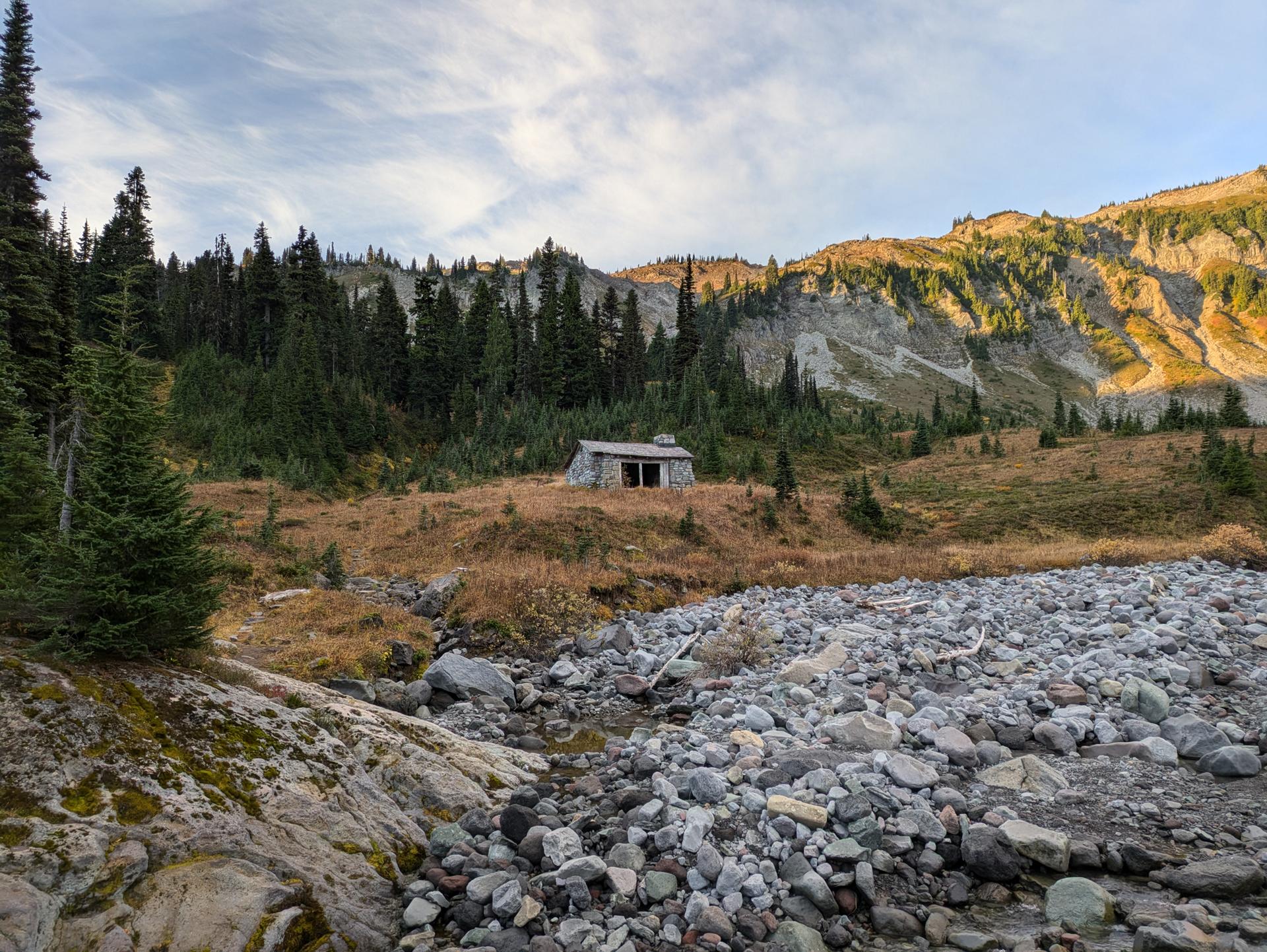 A small rustic cabin in a grassy field with a rocky riverbed and mountains.