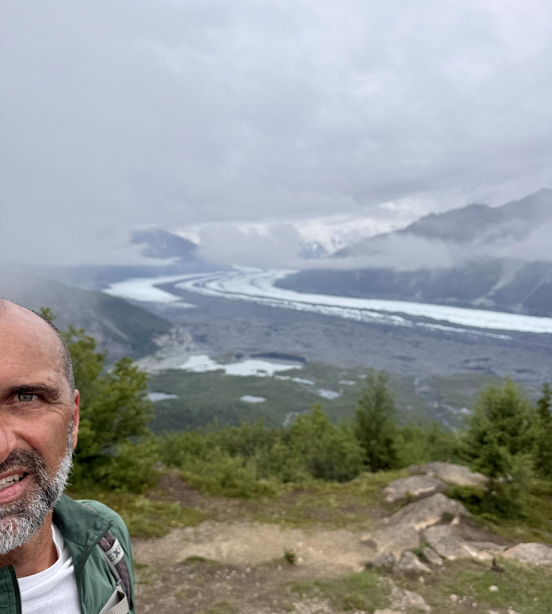 Smiling man's face in foreground, glacier valley and cloudy mountains behind.