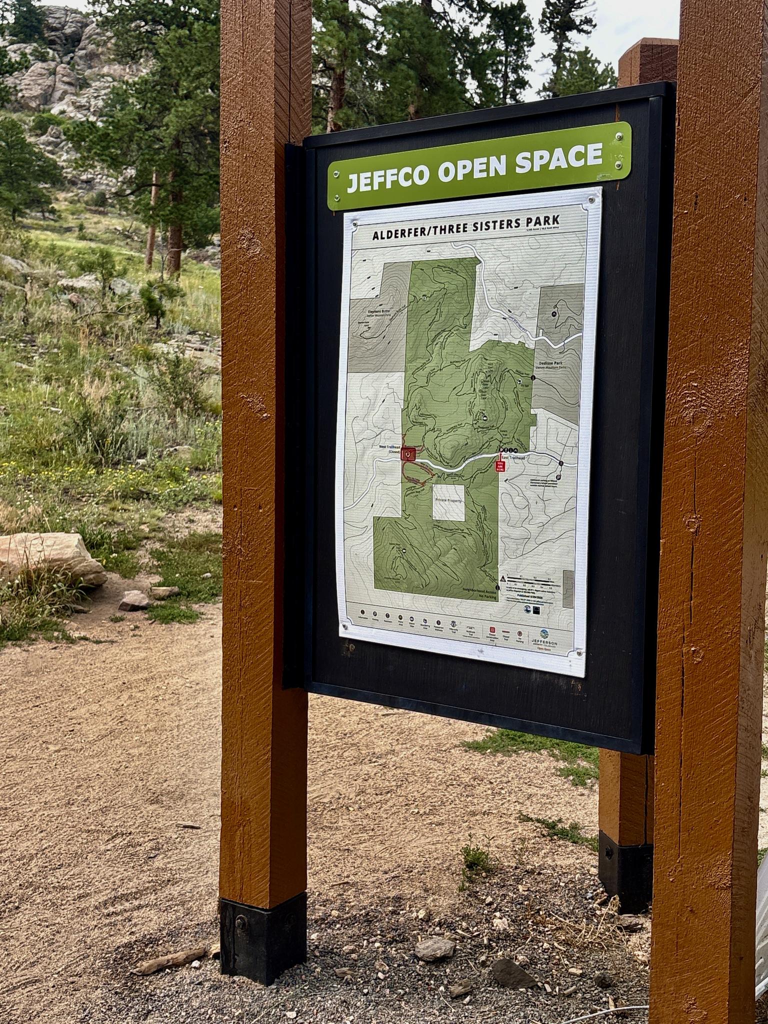 Wooden signpost with a map on a dirt path, surrounded by trees and rocks.