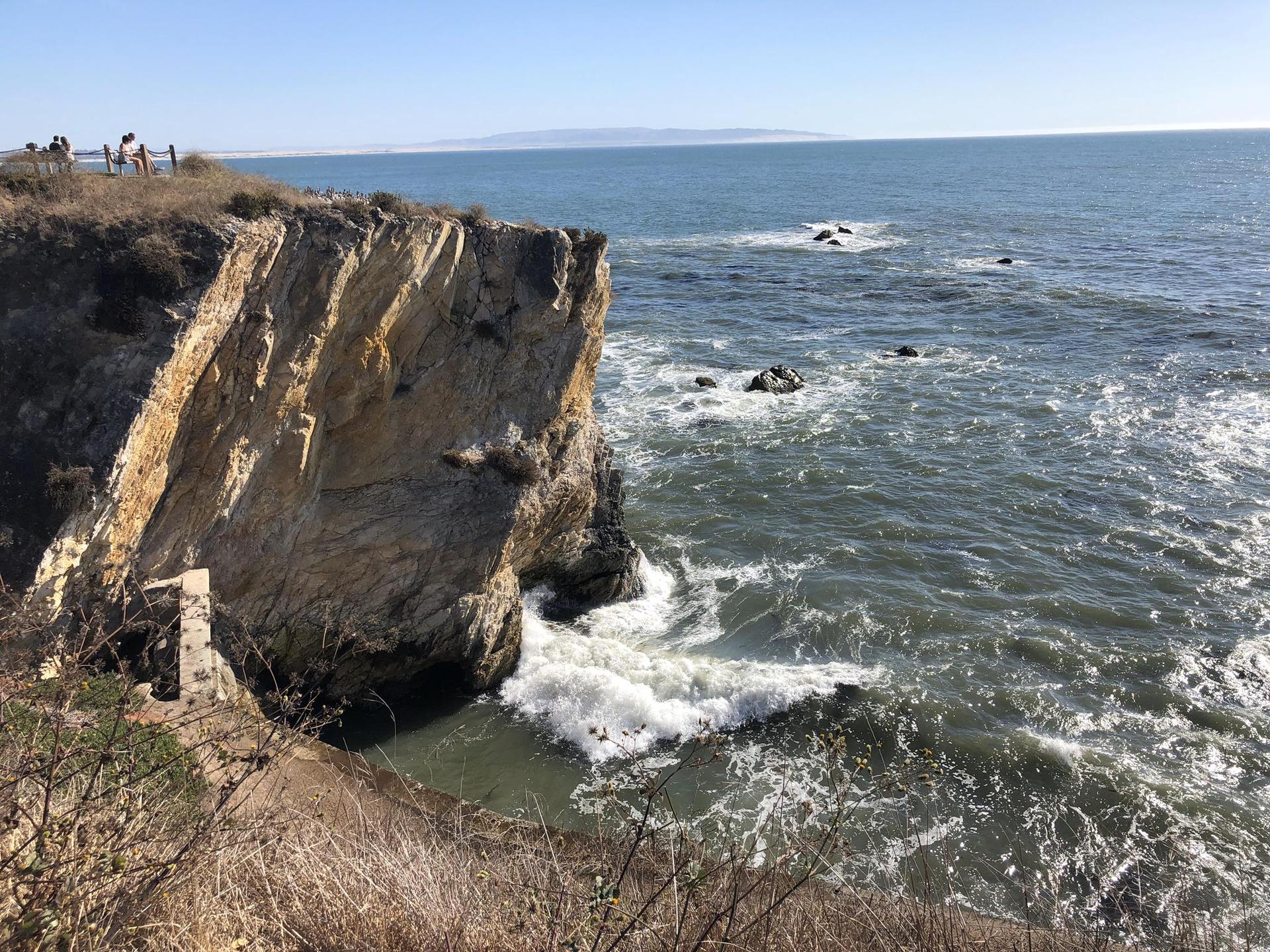 Sunny view of steep ocean cliffs with waves crashing below, distant people.