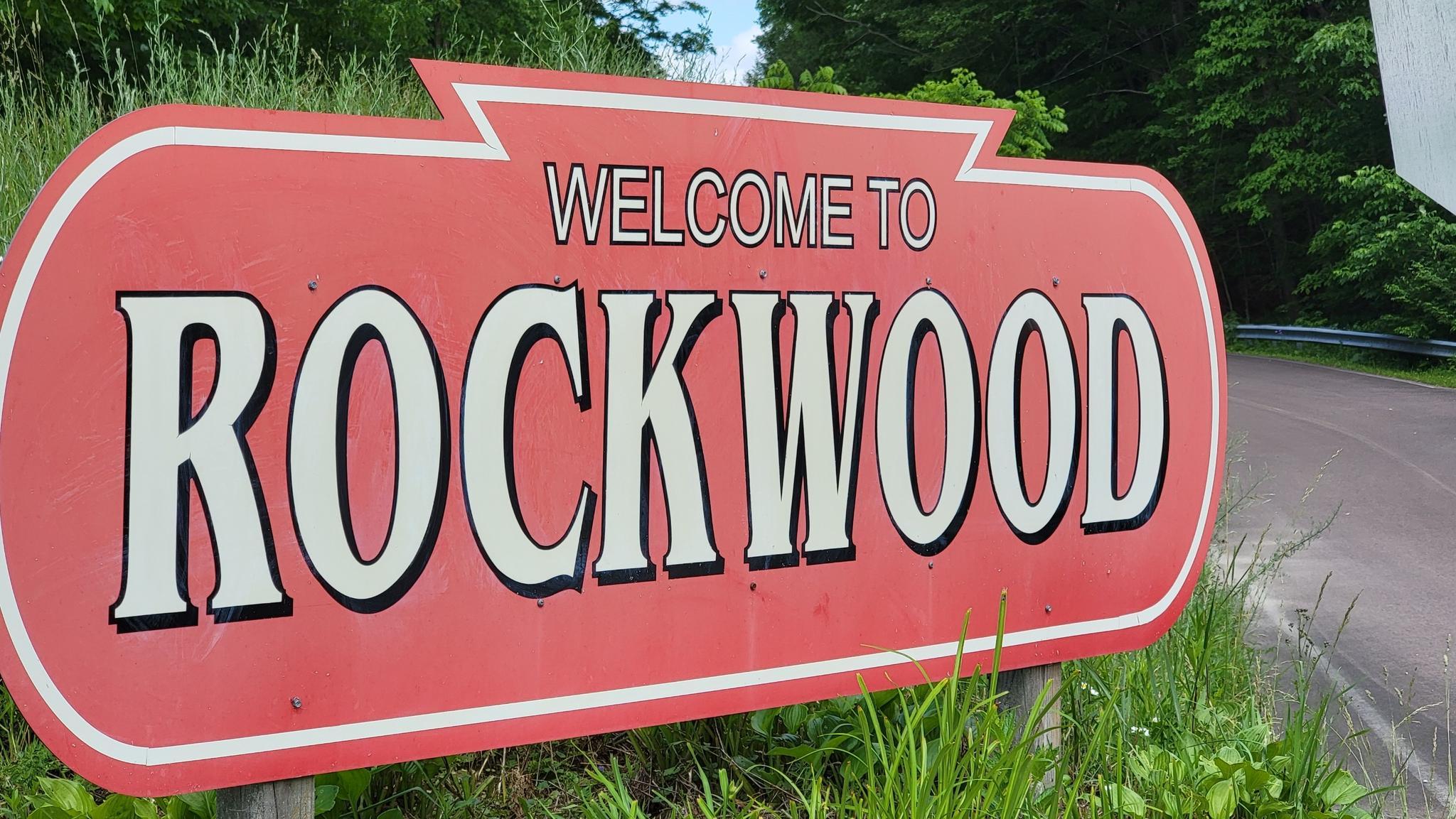 A red and white welcome sign stands by a grassy roadside.