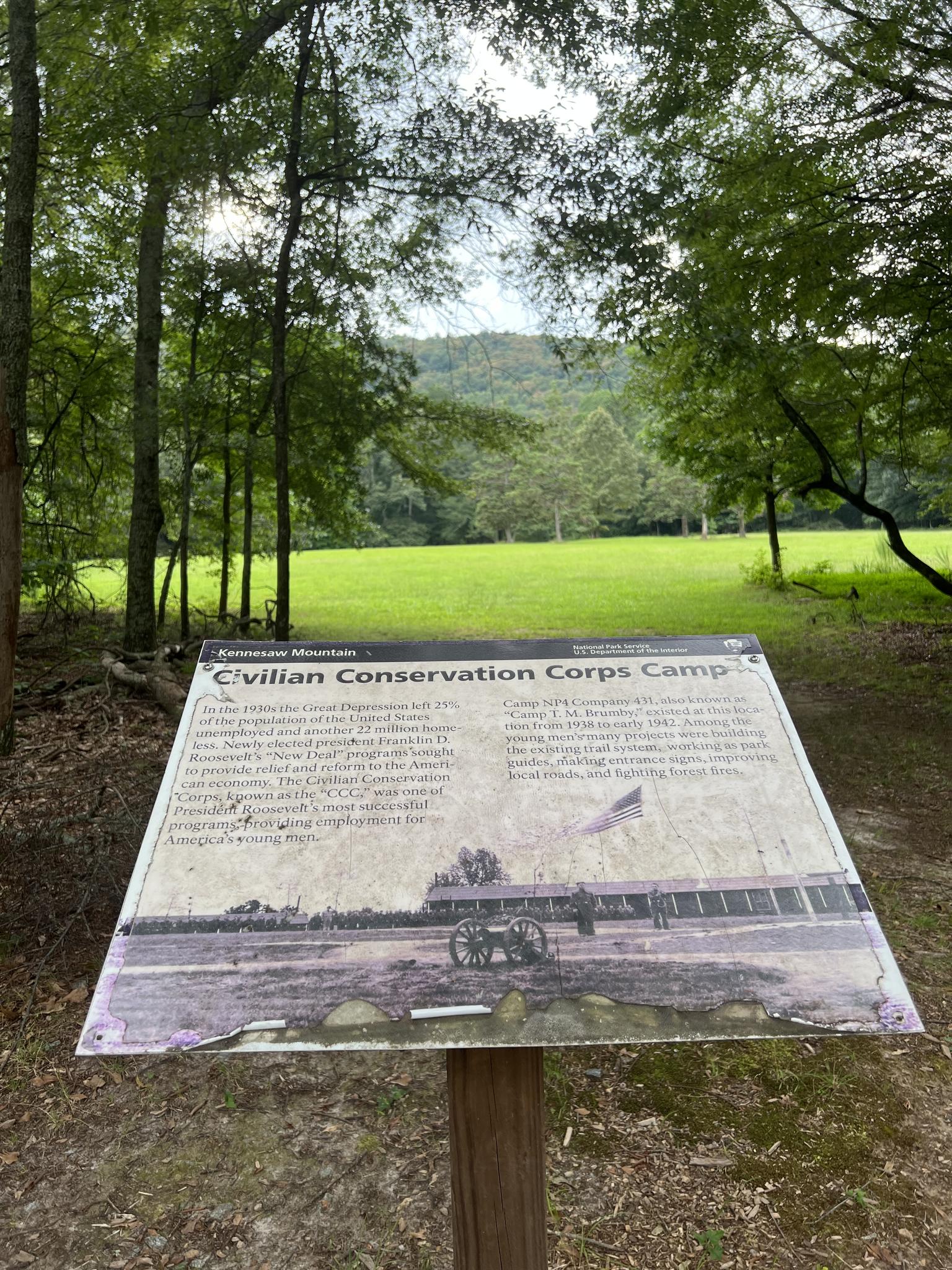 An interpretive sign stands at a forest edge overlooking a green field.